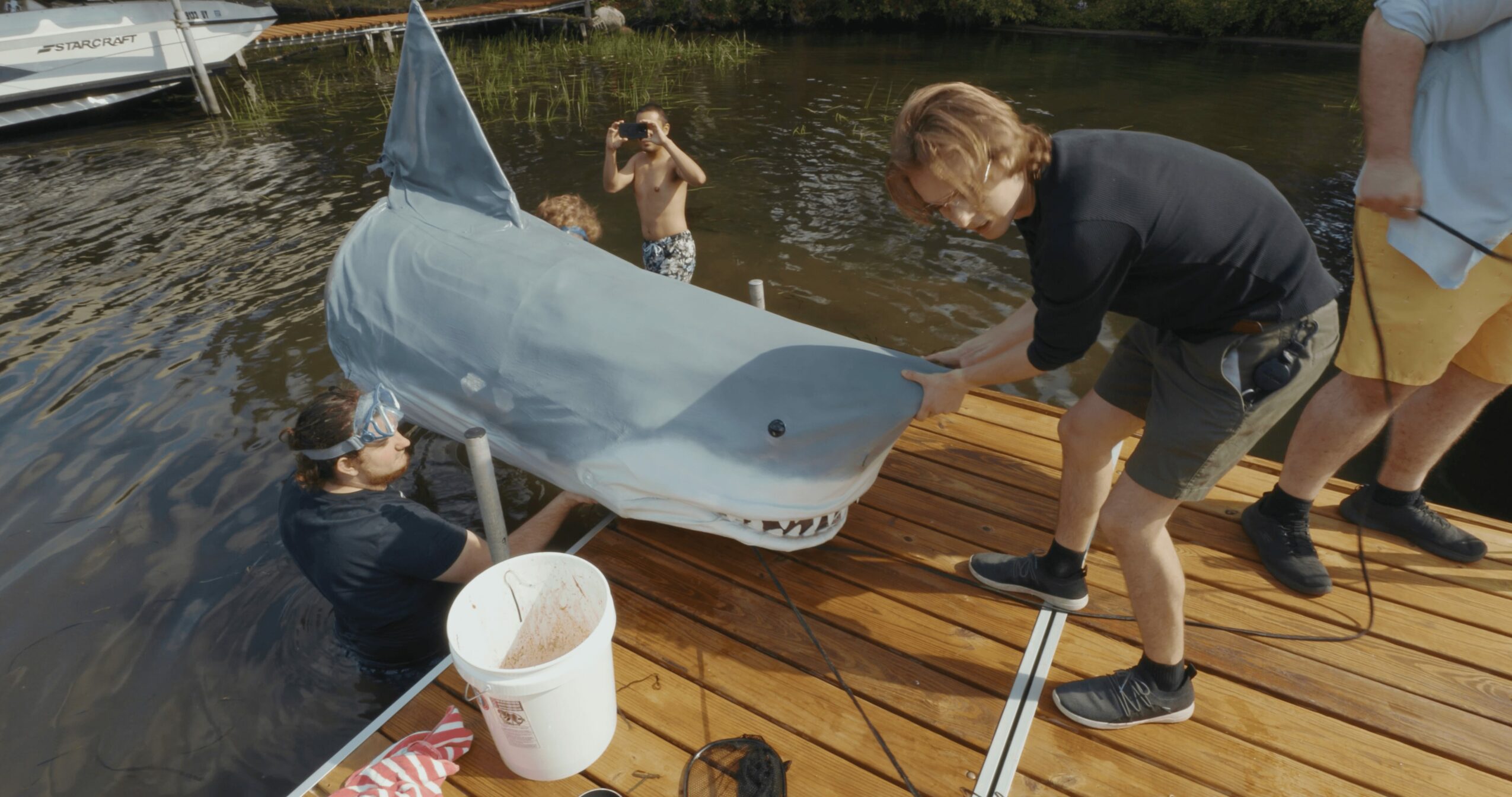 People on a dock with a large shark model, one person holding it, others nearby, water and trees in background.