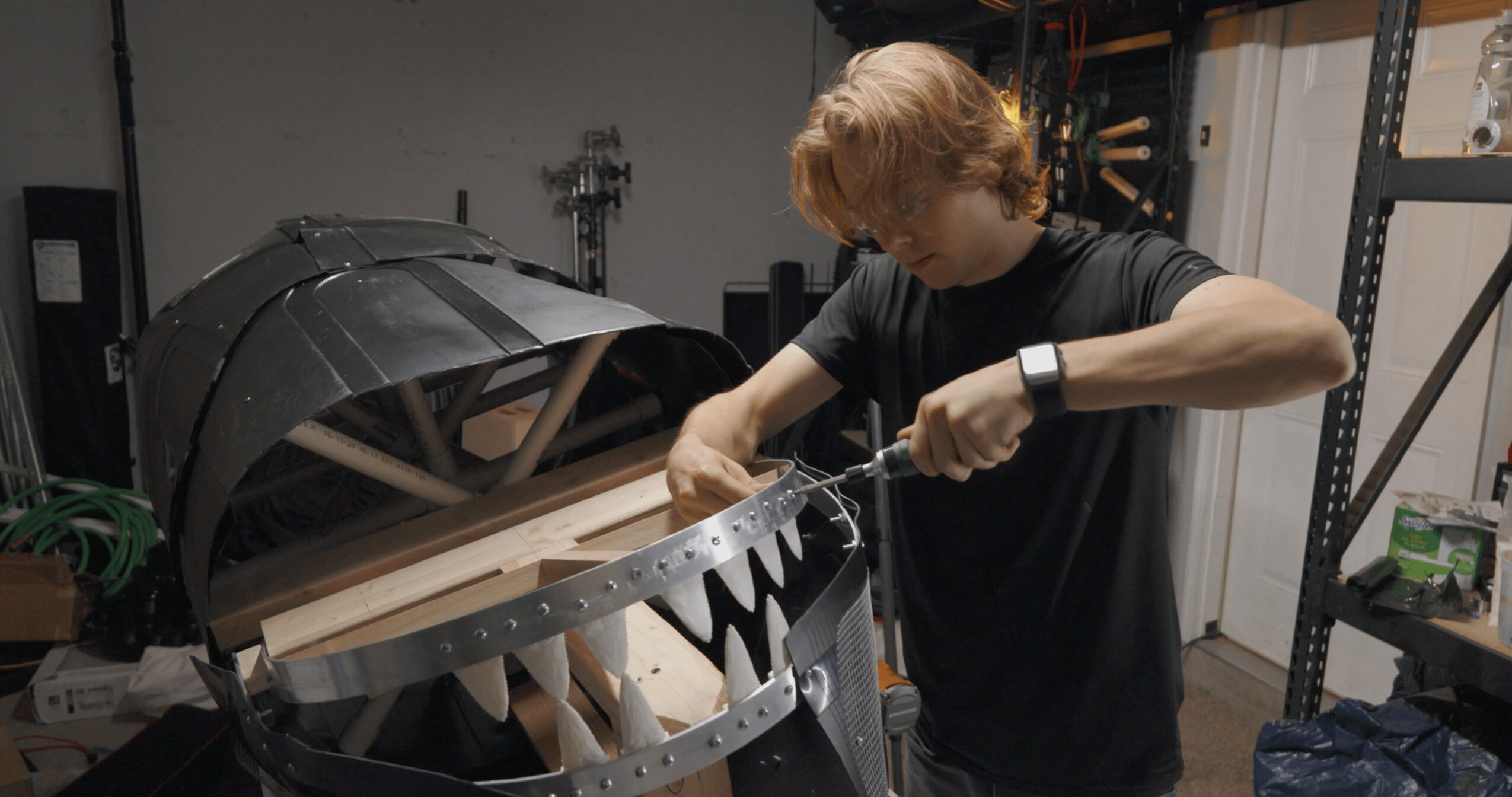 Person working on a large circular mechanical device in a workshop, using a tool, with shelves and equipment nearby.
