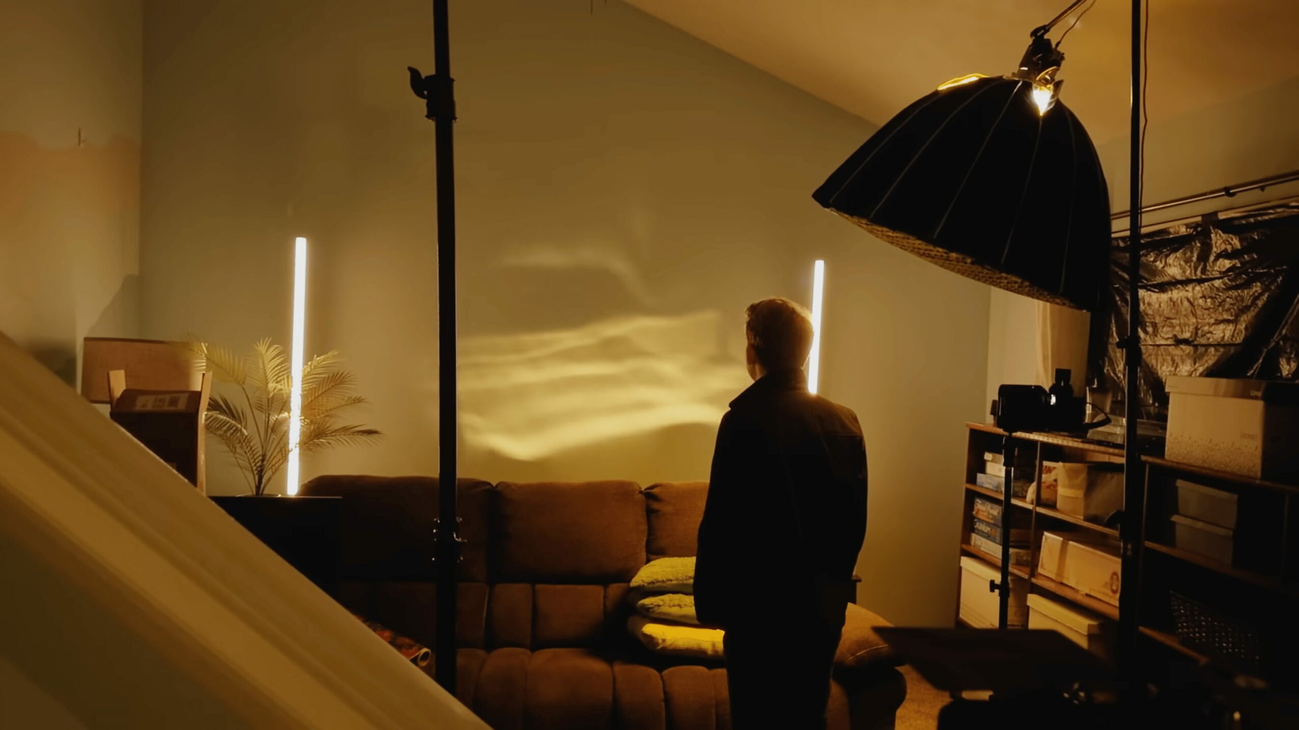 Person standing in a dimly lit room with a sofa, large lamp, and shelves, facing away from the camera.