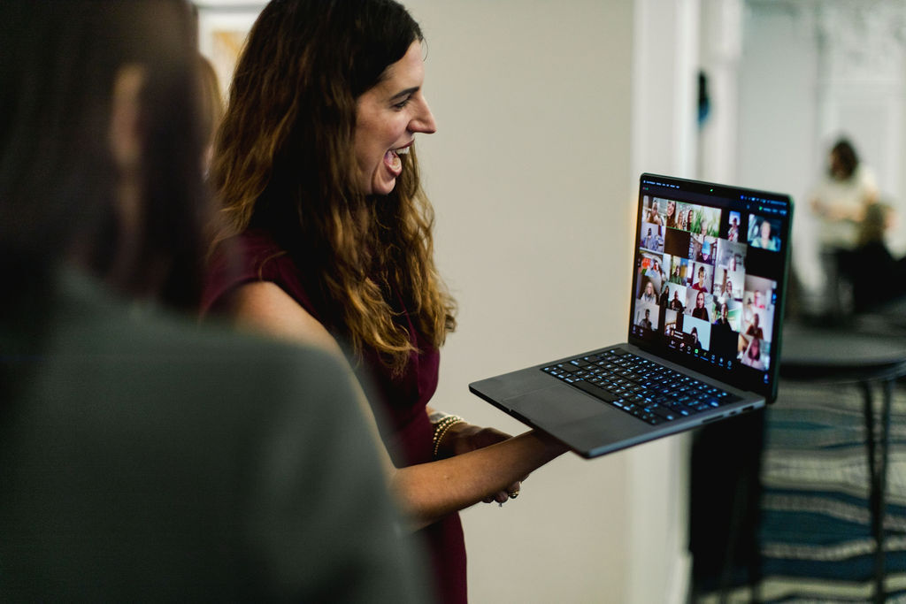 Woman smiling and holding a laptop with a video conference screen, in a room with a blurred background.