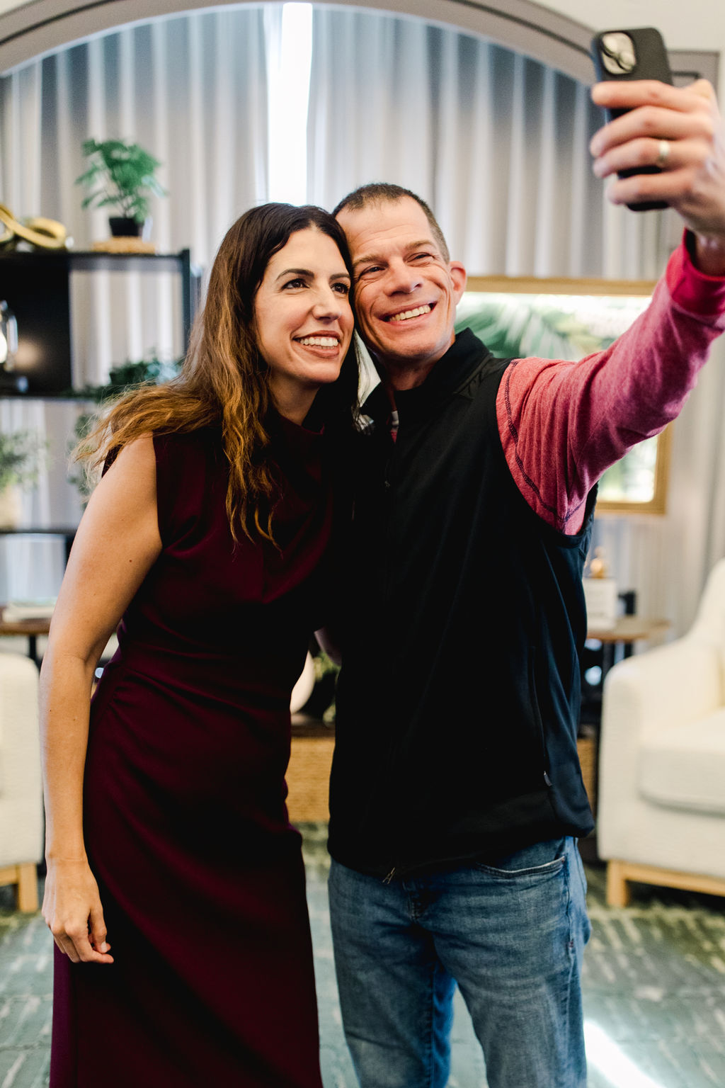 Two people taking a selfie indoors, smiling, with a woman in a dark dress and a man in a dark vest and jeans, holding a phone.