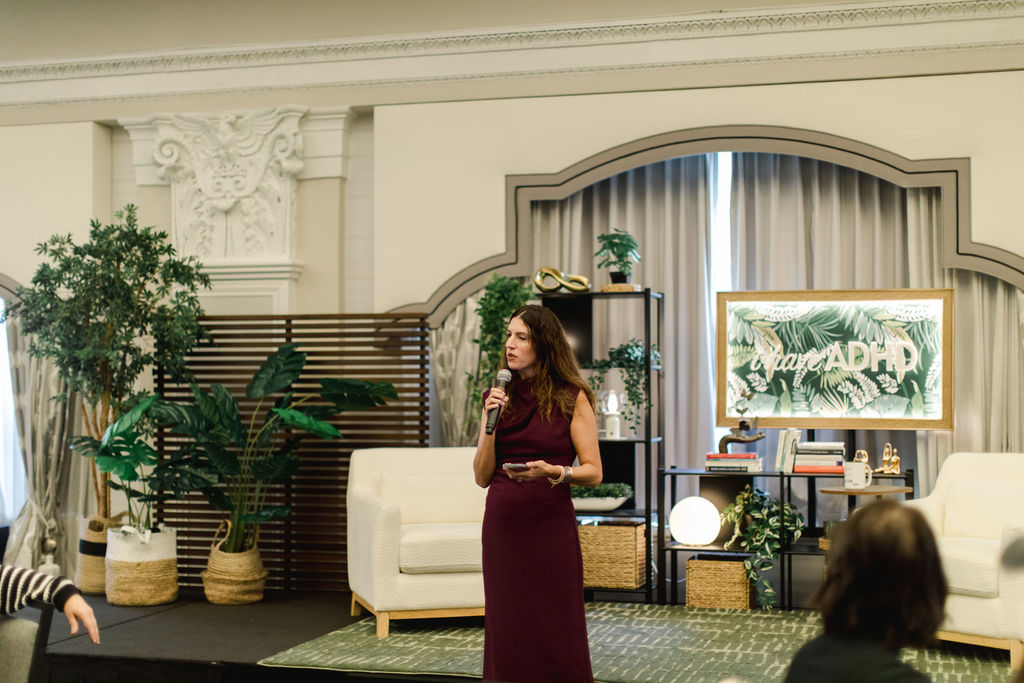 Woman in a burgundy dress speaking into a microphone on a stage with plants and furniture, audience visible in foreground.