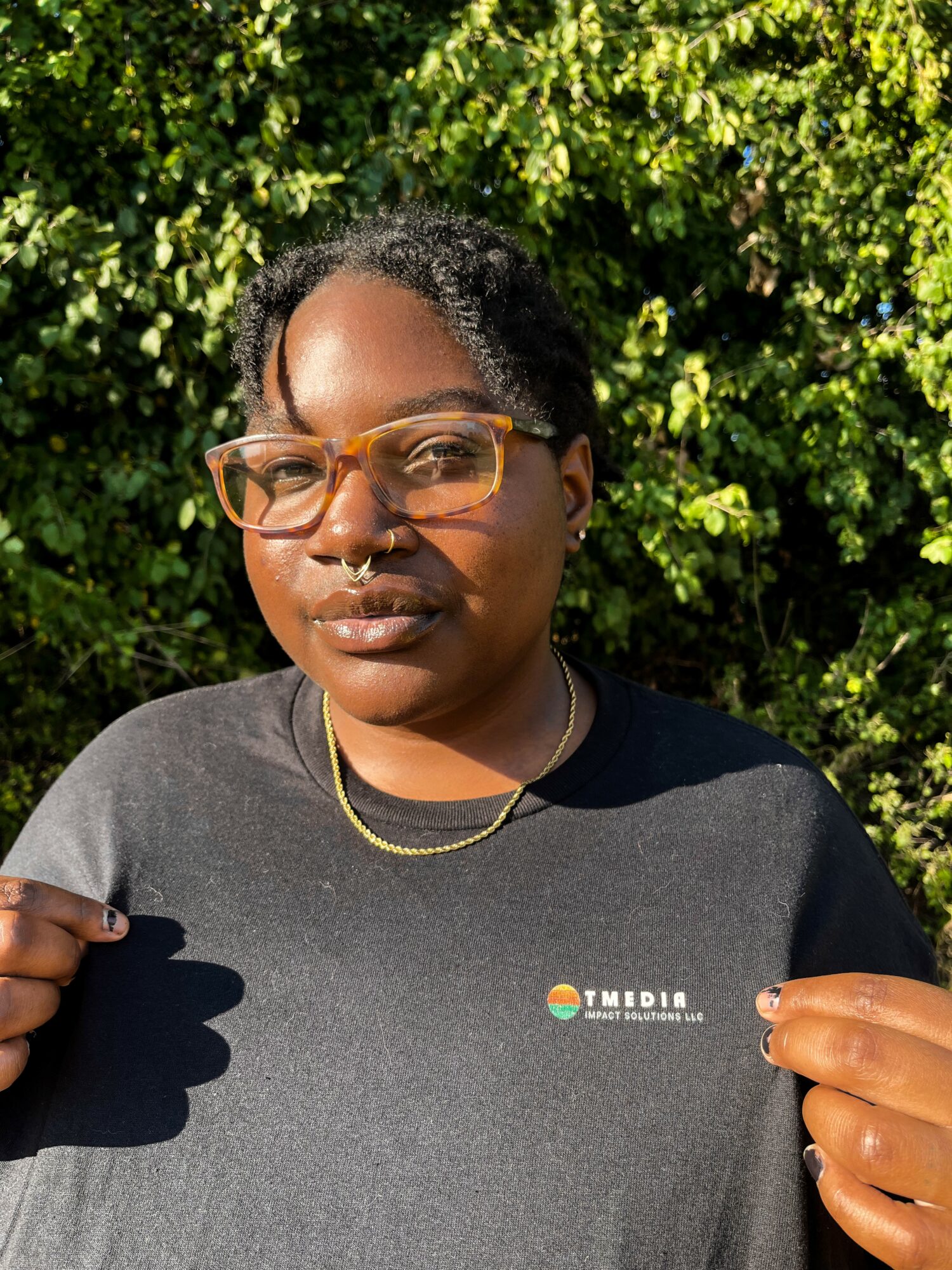 Woman with glasses and a black T-shirt outdoors, green foliage background, sunlight on face, holding the shirt's collar.