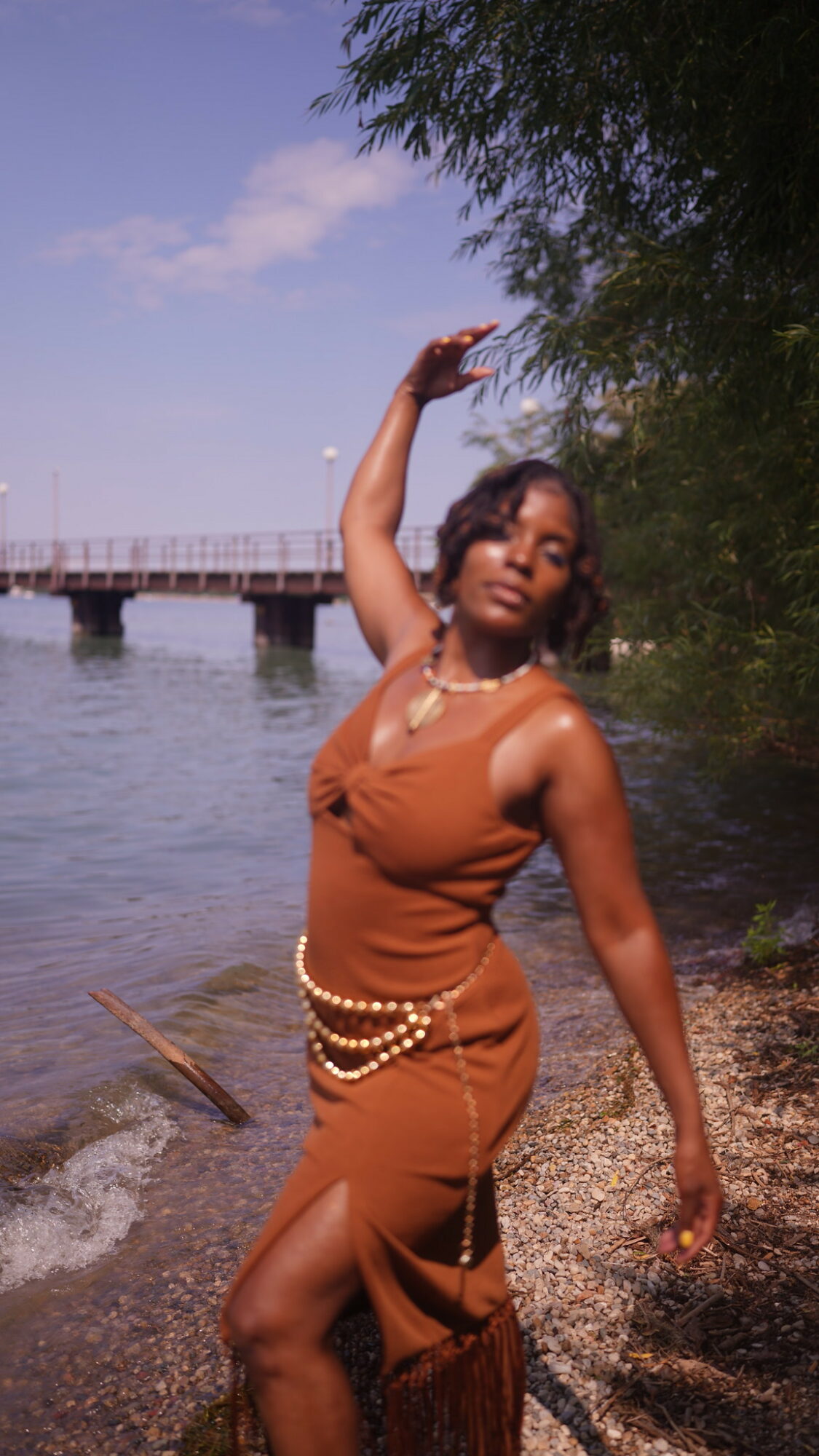 Woman in brown dress standing on a beach near water with trees and a pier in background.