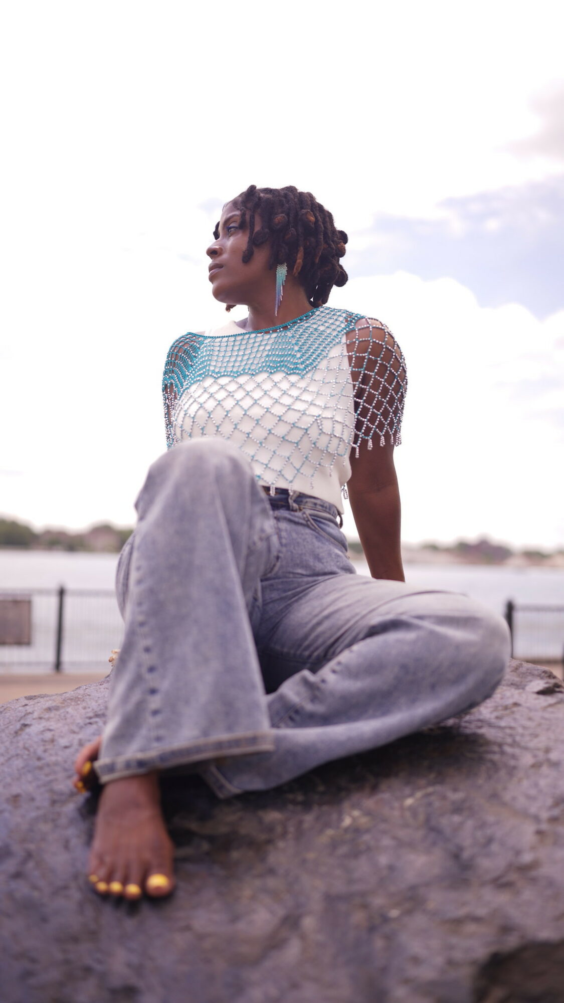 Woman with short curly hair sitting outdoors on a rock, wearing a white and blue top and light jeans, looking to the side.