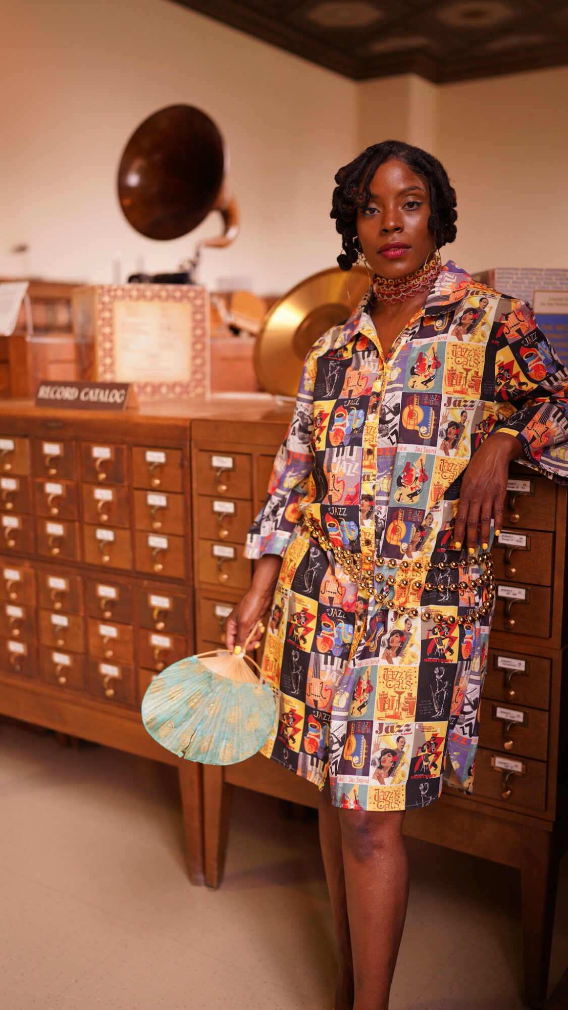 Woman in colorful patterned coat standing next to a wooden cabinet with vintage records and a hat, holding a striped bag.