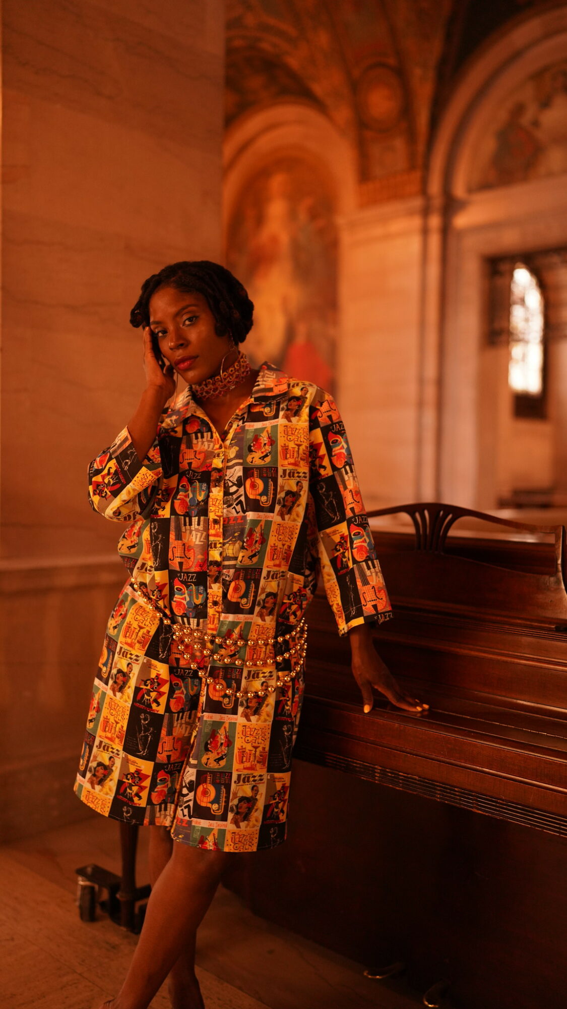 Woman in colorful patterned dress standing near a piano inside a grand hall with arches and artwork.