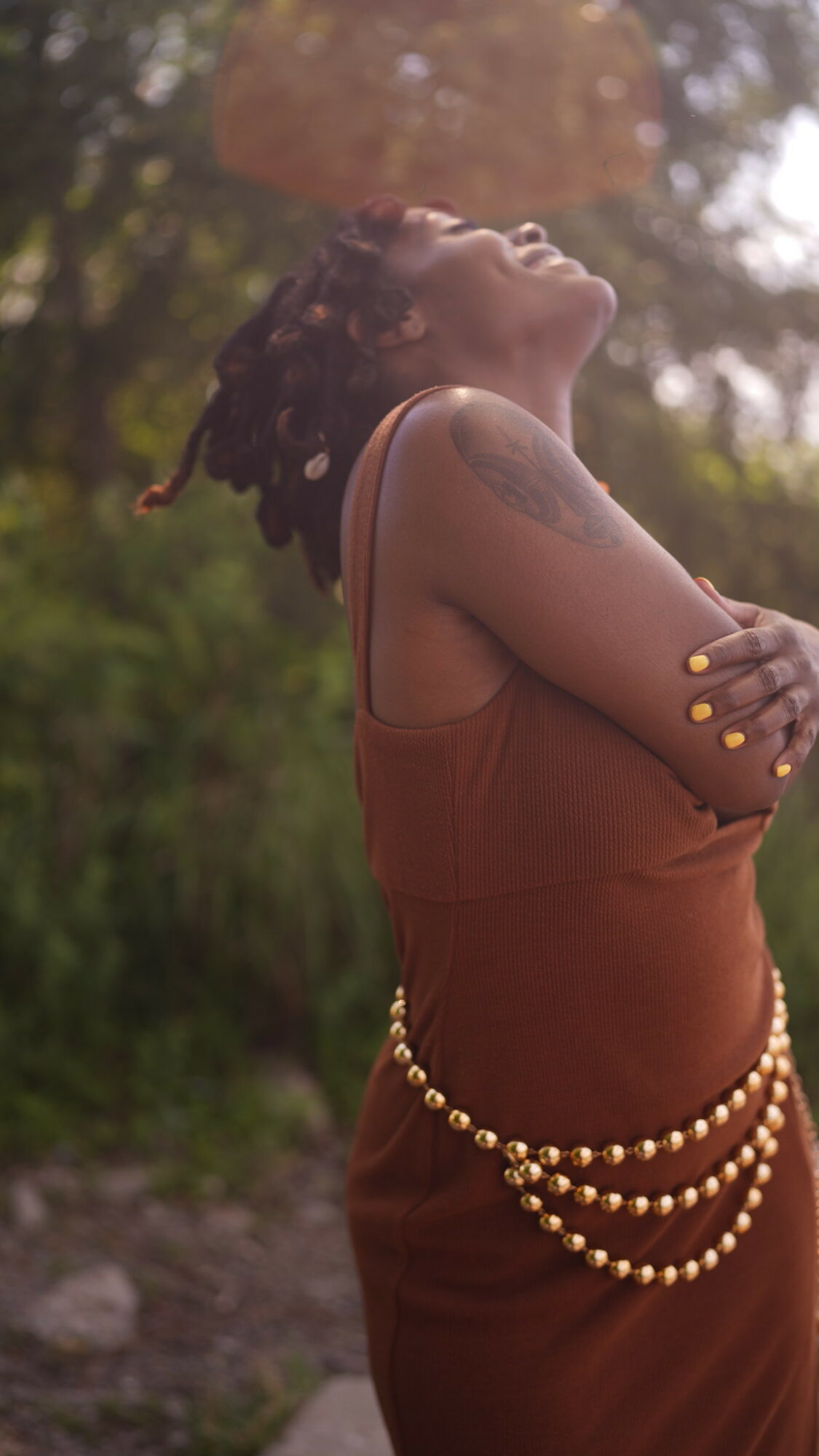 Woman with dreadlocks smiling outdoors, wearing a brown dress and pearl accessories, with sunlight filtering through trees.