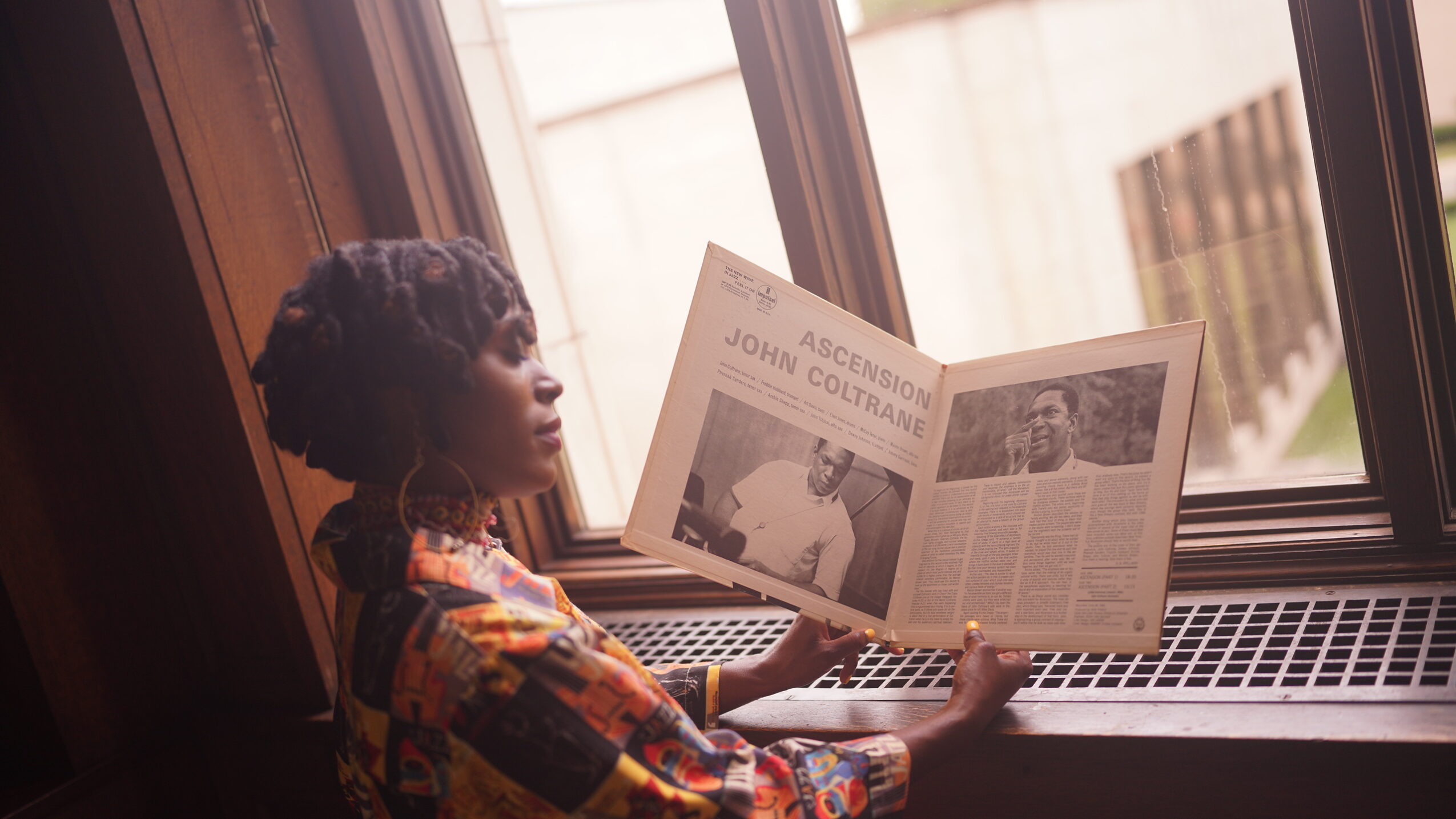 Person with curly hair reading a newspaper by a window, natural light illuminating the scene.
