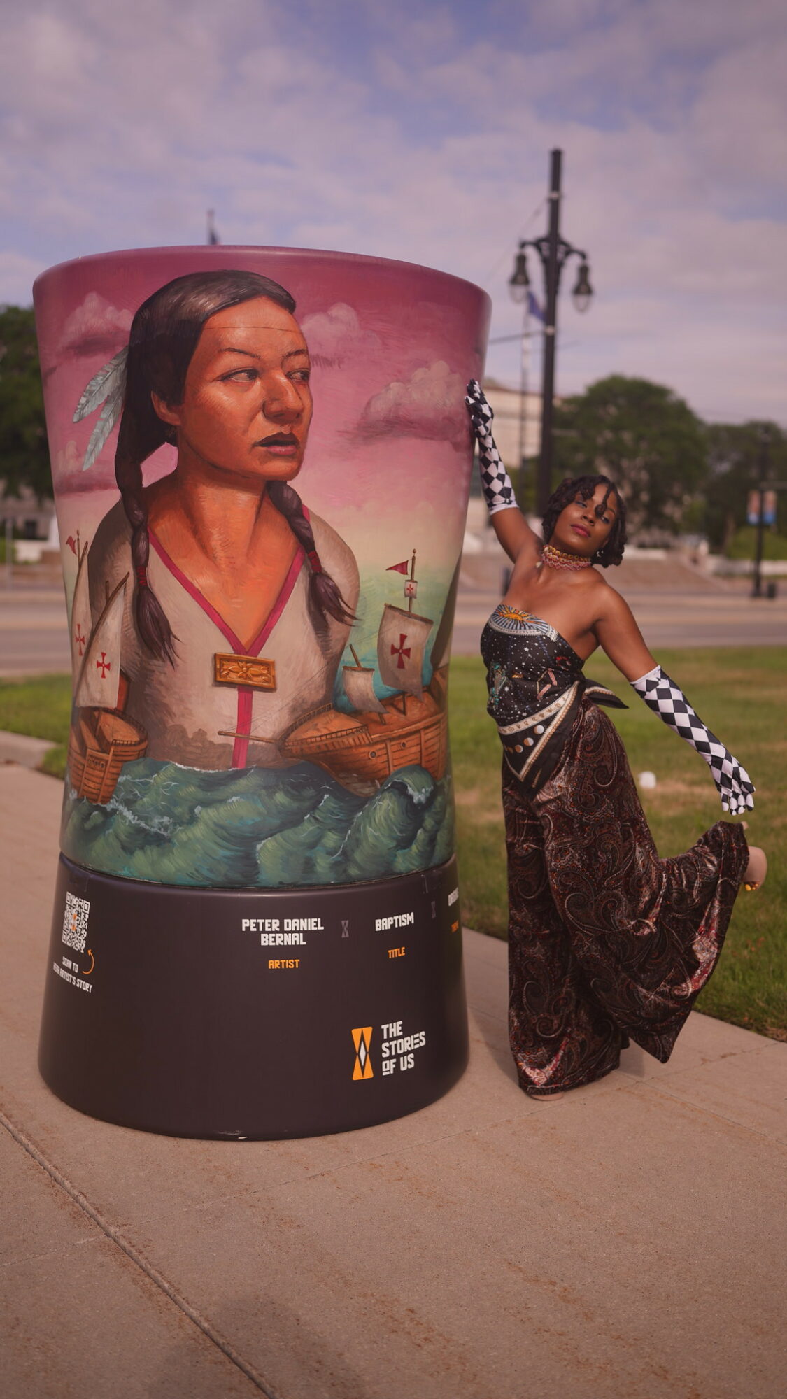 Person in traditional attire holding a flag next to a large painted cylindrical sculpture of a woman with a landscape background.