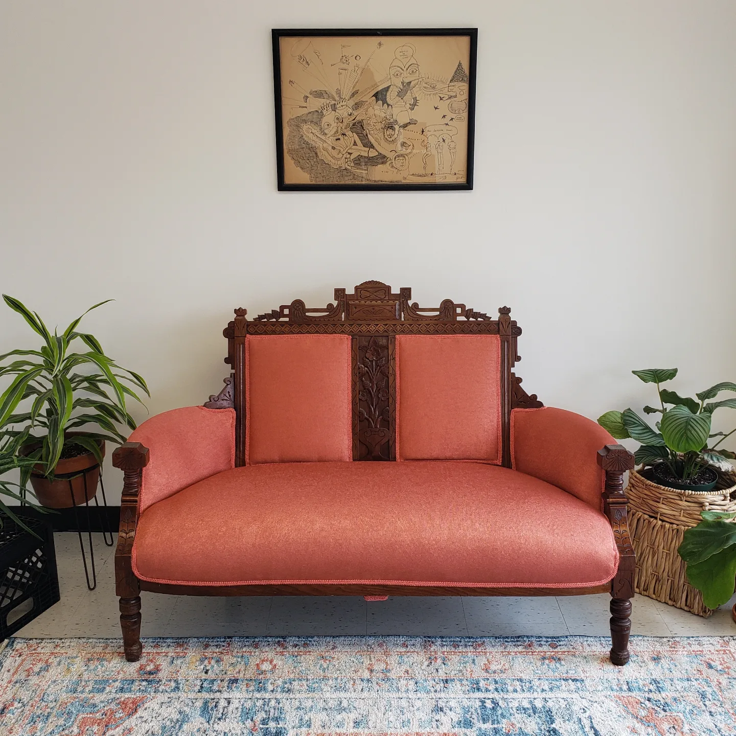 Vintage pink upholstered sofa with wooden frame, two plants on sides, framed artwork above, and a patterned rug below.