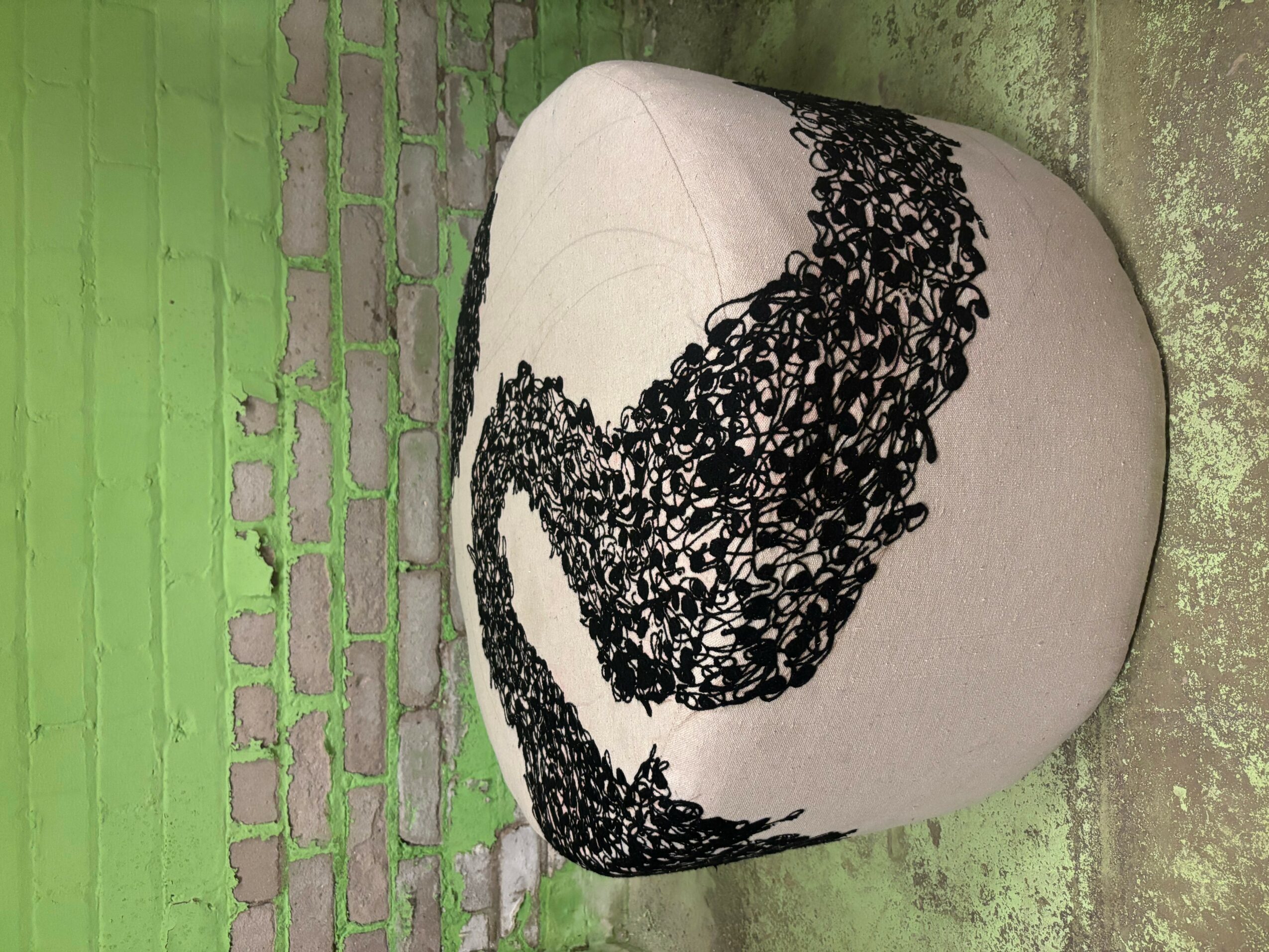Upright beige ottoman with black lace pattern, against a green brick wall and concrete floor.