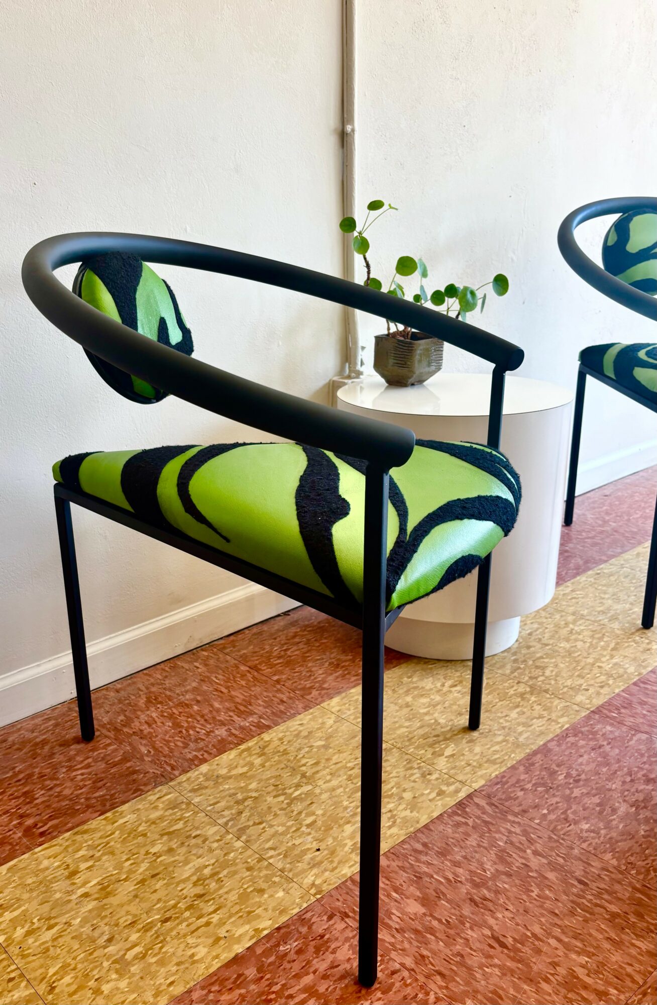 Chair with black frame and green and black patterned cushion, next to a small table with a plant, on a multicolored floor.