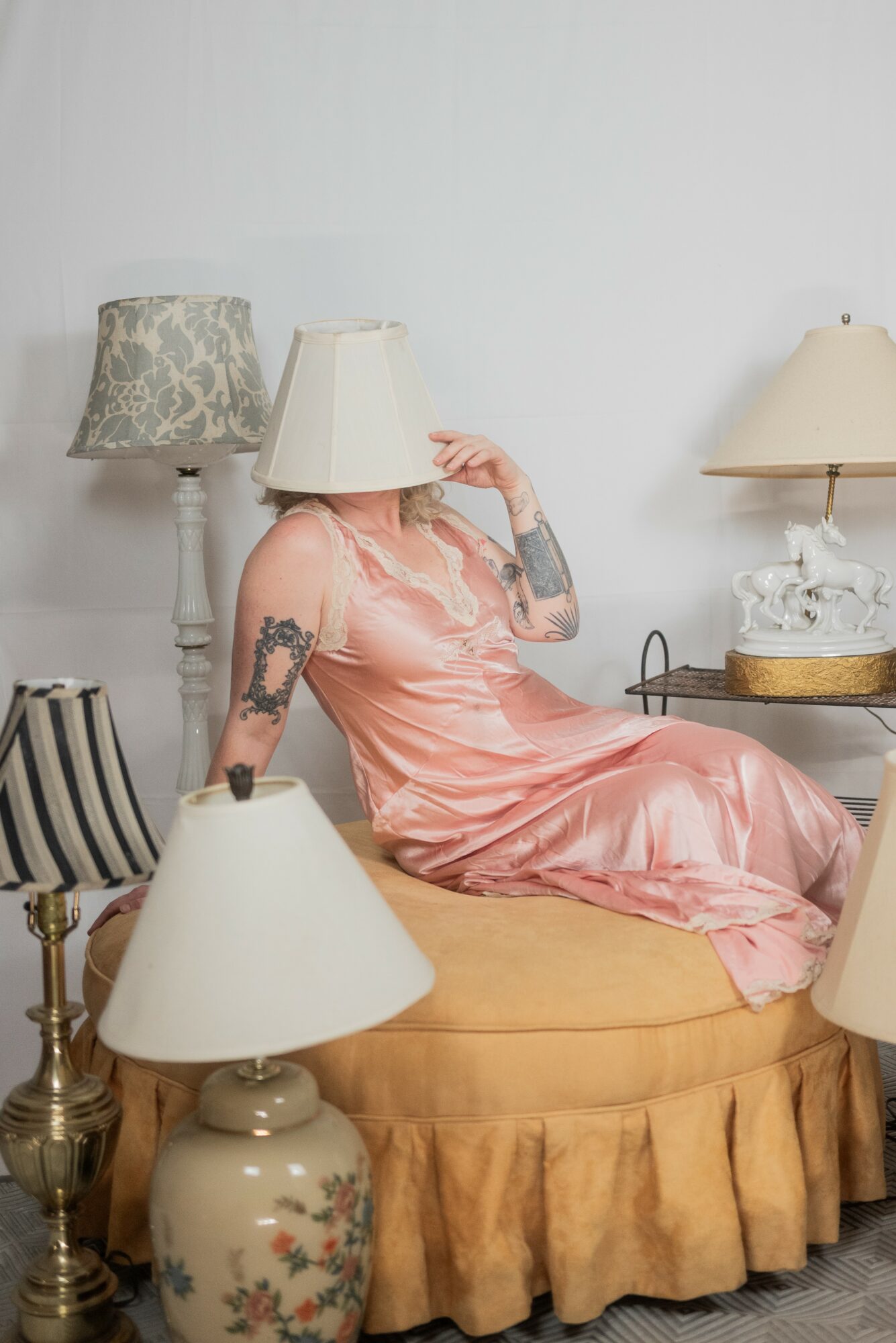 Woman in pink dress sitting on a yellow ottoman, surrounded by lamps and decorative items, with her face obscured by a lampshade.