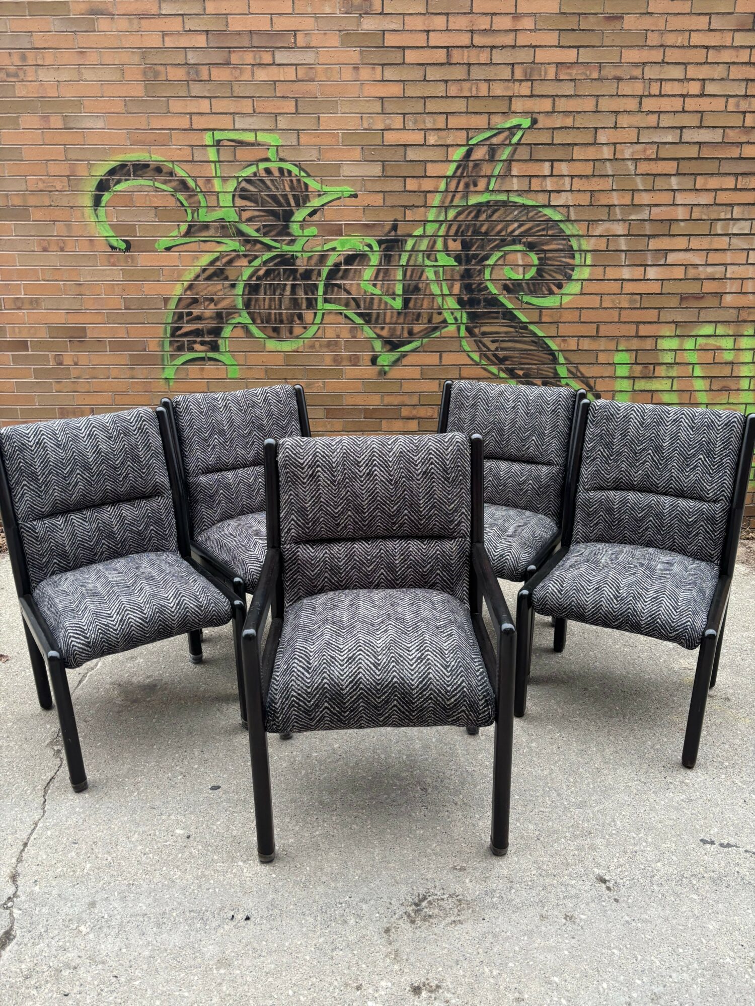 Six gray upholstered chairs with black metal legs arranged in two rows on a concrete floor, brick wall background, and a green vine plant.