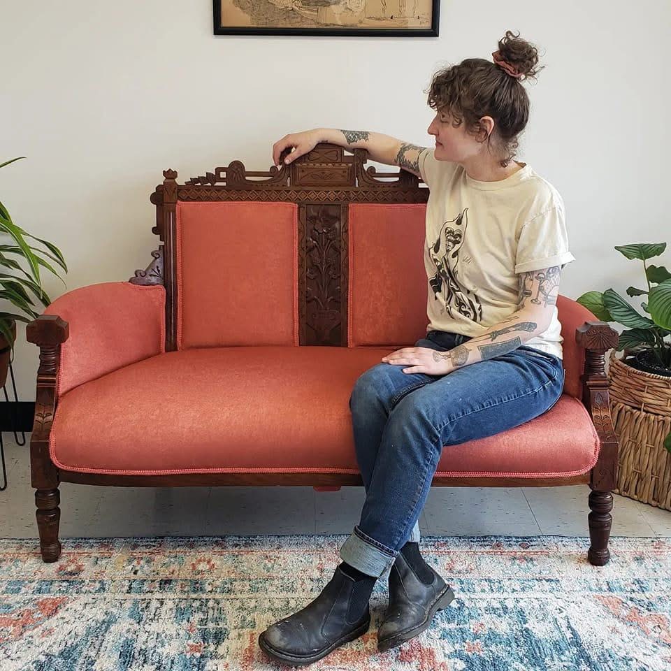 Young woman with tattoos sitting on a vintage pink sofa, looking to the side, in a room with plants and a rug.