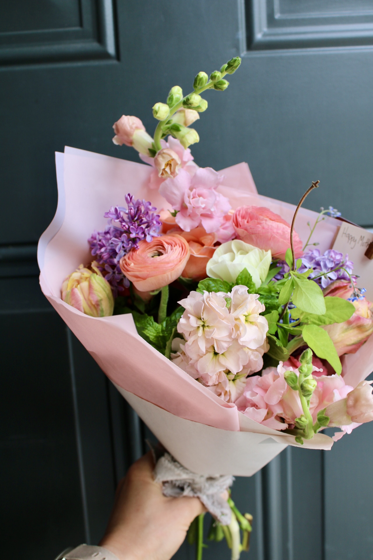 Hand holding a colorful bouquet of pink, purple, and white flowers wrapped in pink paper against a dark background.
