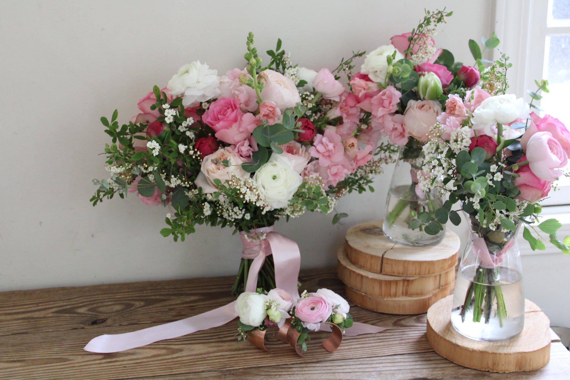 Two floral arrangements with pink and white flowers, greenery, and pink ribbons on wooden and glass bases, on a wooden surface.