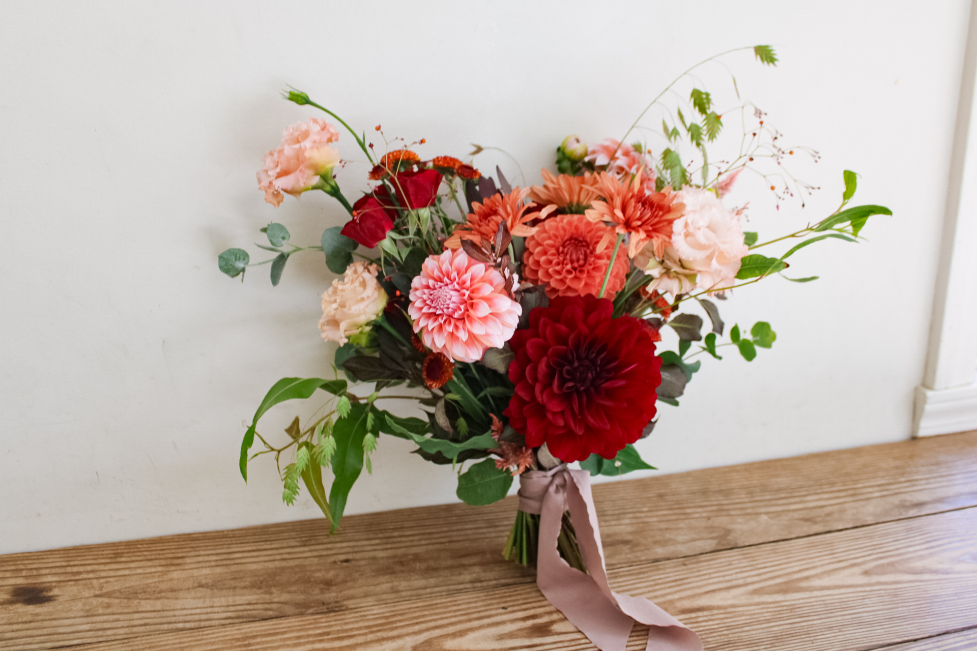 Bouquet of pink, red, and orange flowers with green leaves, tied with a pink ribbon, on a wooden surface.