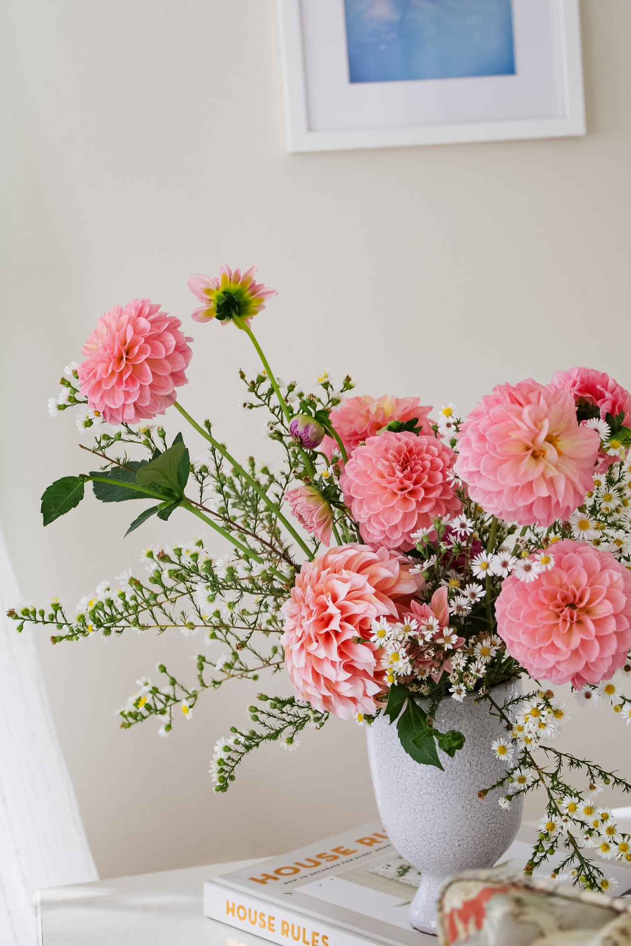 Pink and white flowers in a white vase on a table, with a framed picture on the wall above.