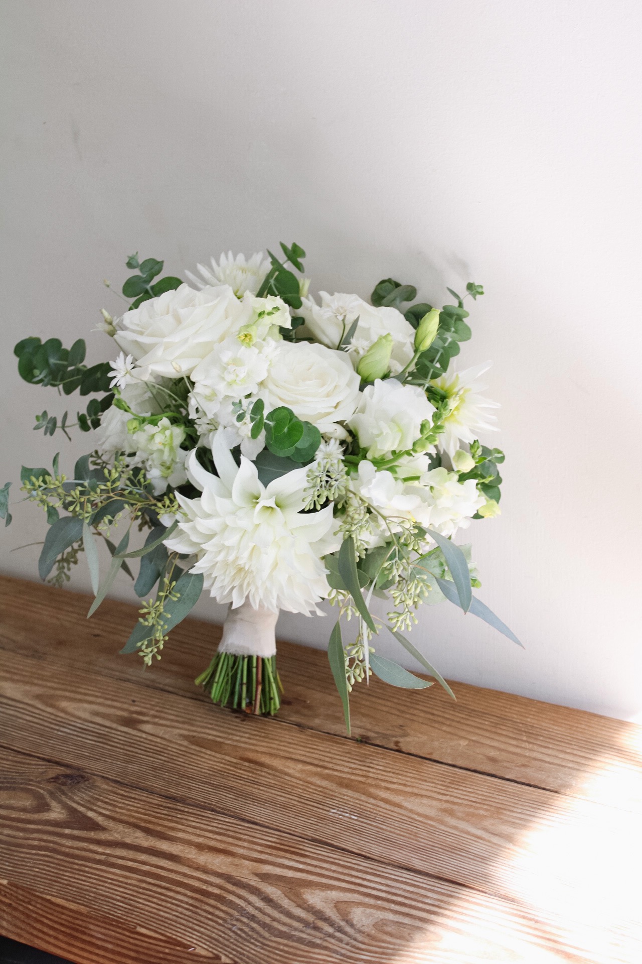 White flower bouquet with greenery on a wooden surface against a plain background.