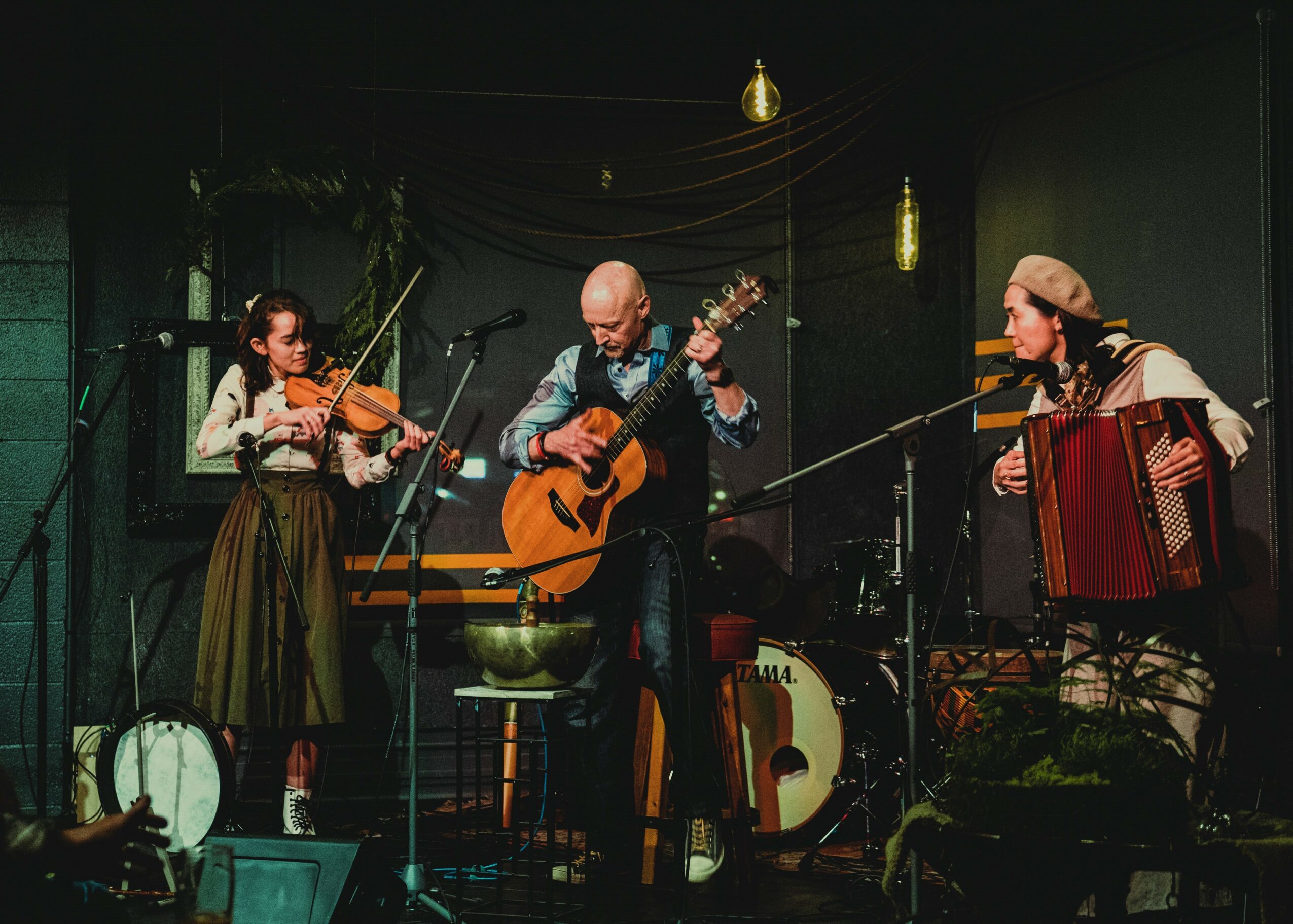Three musicians perform on stage with instruments, including a violin, guitar, and accordion, in a dimly lit setting.