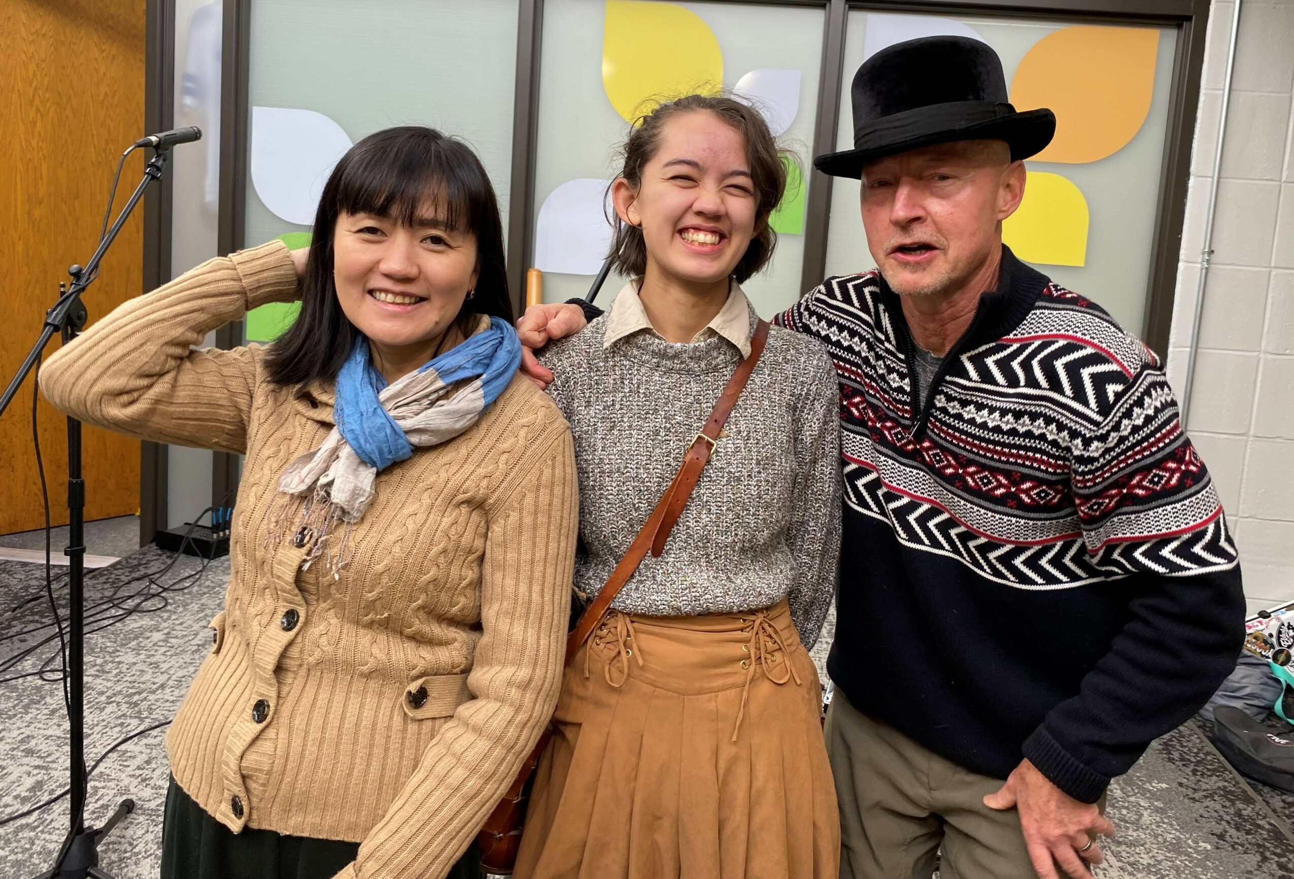 Three people smiling, standing close together indoors, with colorful background panels, one wearing a hat, two women with scarves.