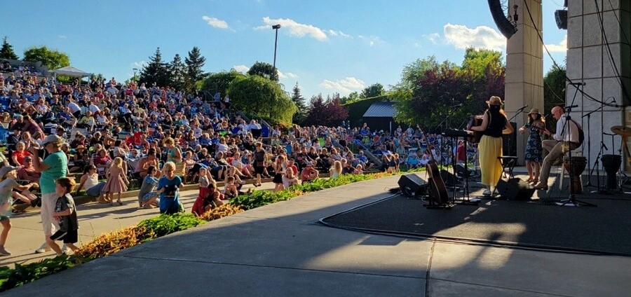 Outdoor concert with a large audience seated on steps, band performing on stage, trees and blue sky in background.