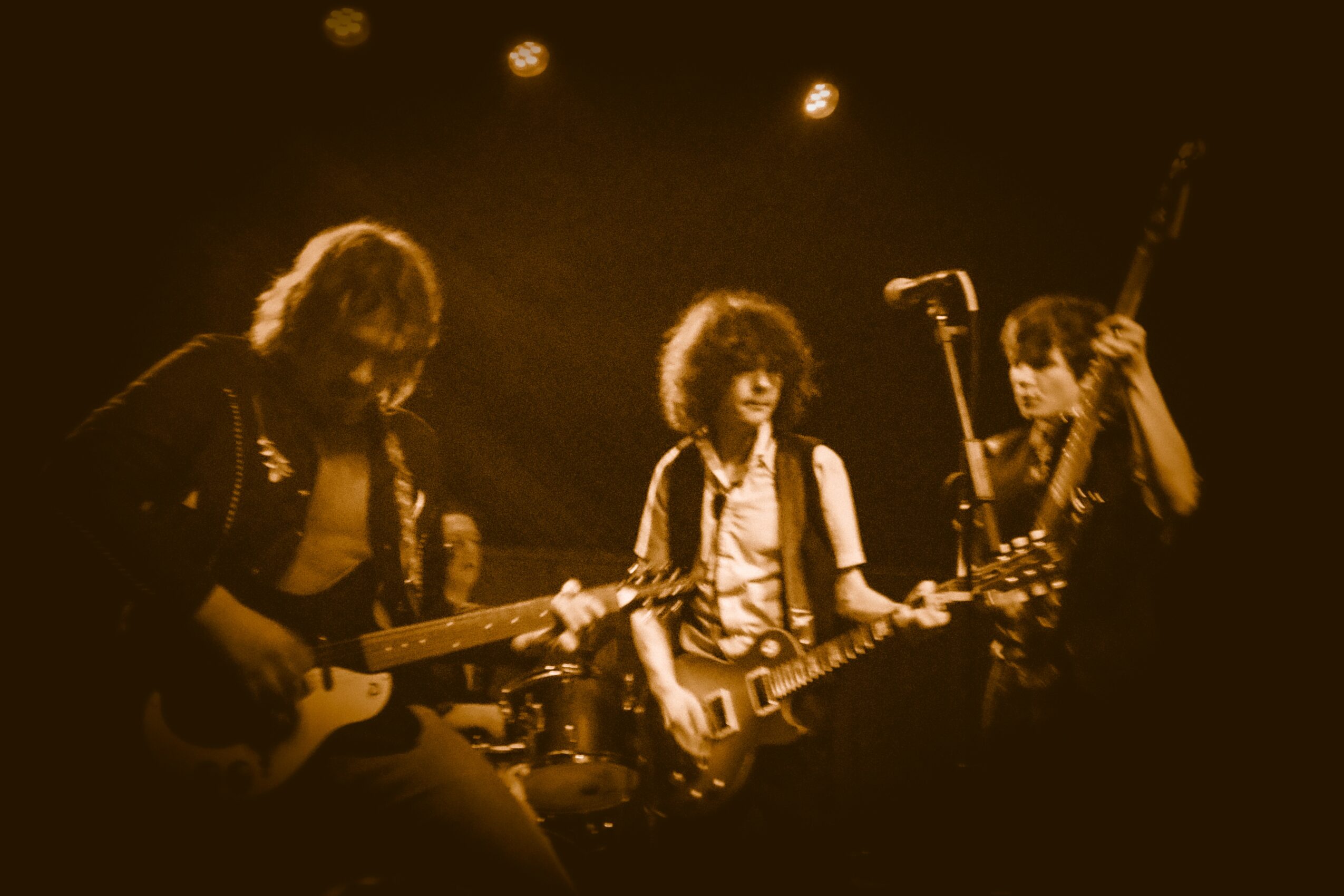 Three musicians playing guitars on stage with warm lighting, one person singing into a microphone, in a live performance setting.