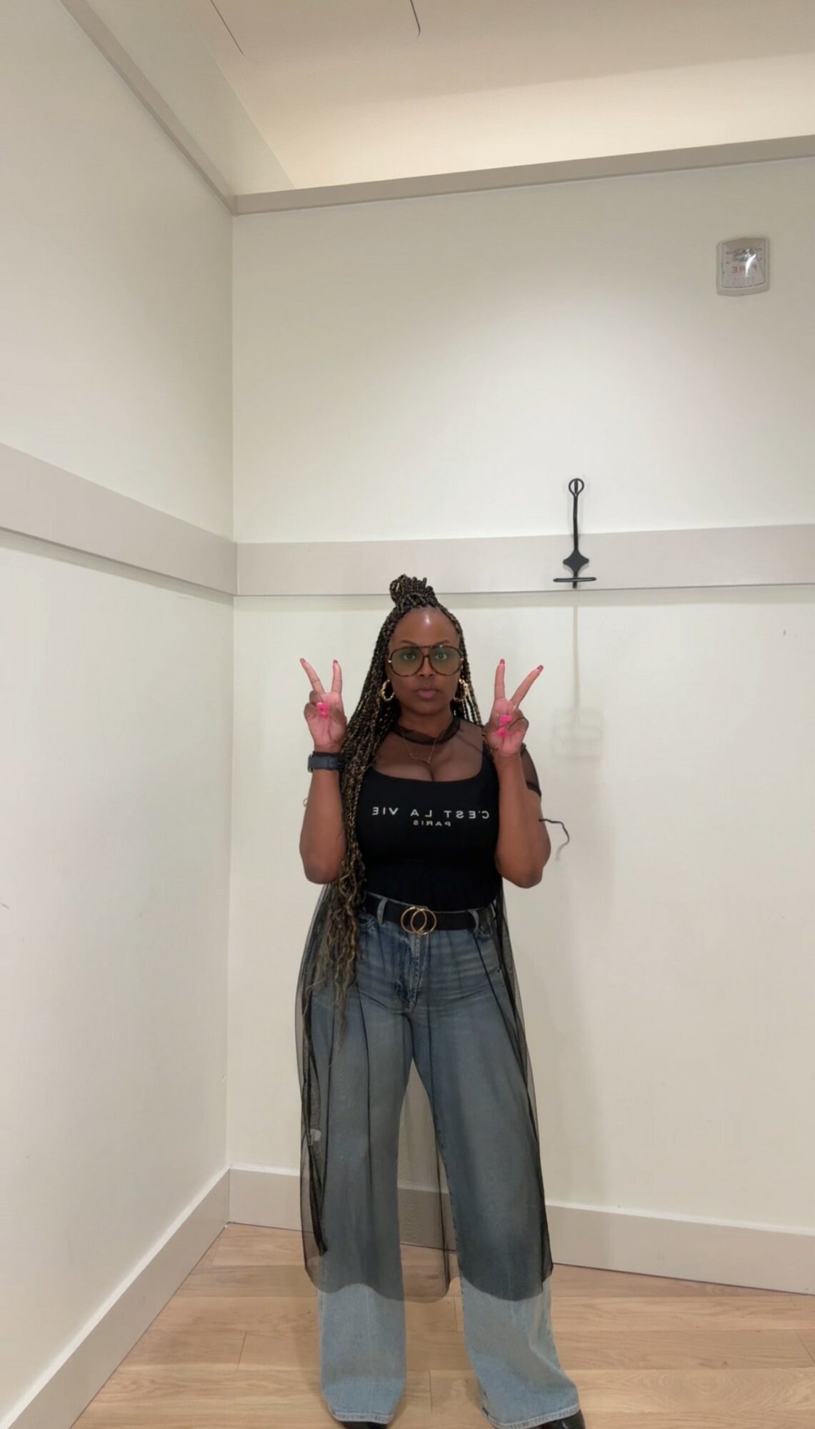 Person standing against a white wall, making peace signs with both hands, wearing glasses, a black top, and wide-leg jeans.