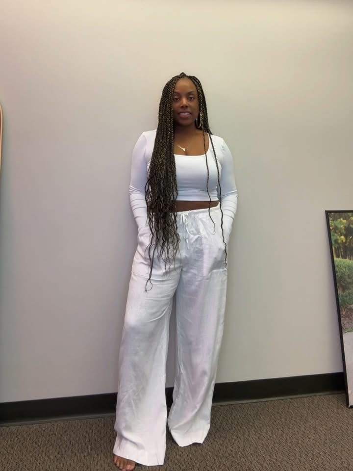 Woman with long braids in white outfit standing against a plain wall, smiling, with a framed picture partially visible on the right.
