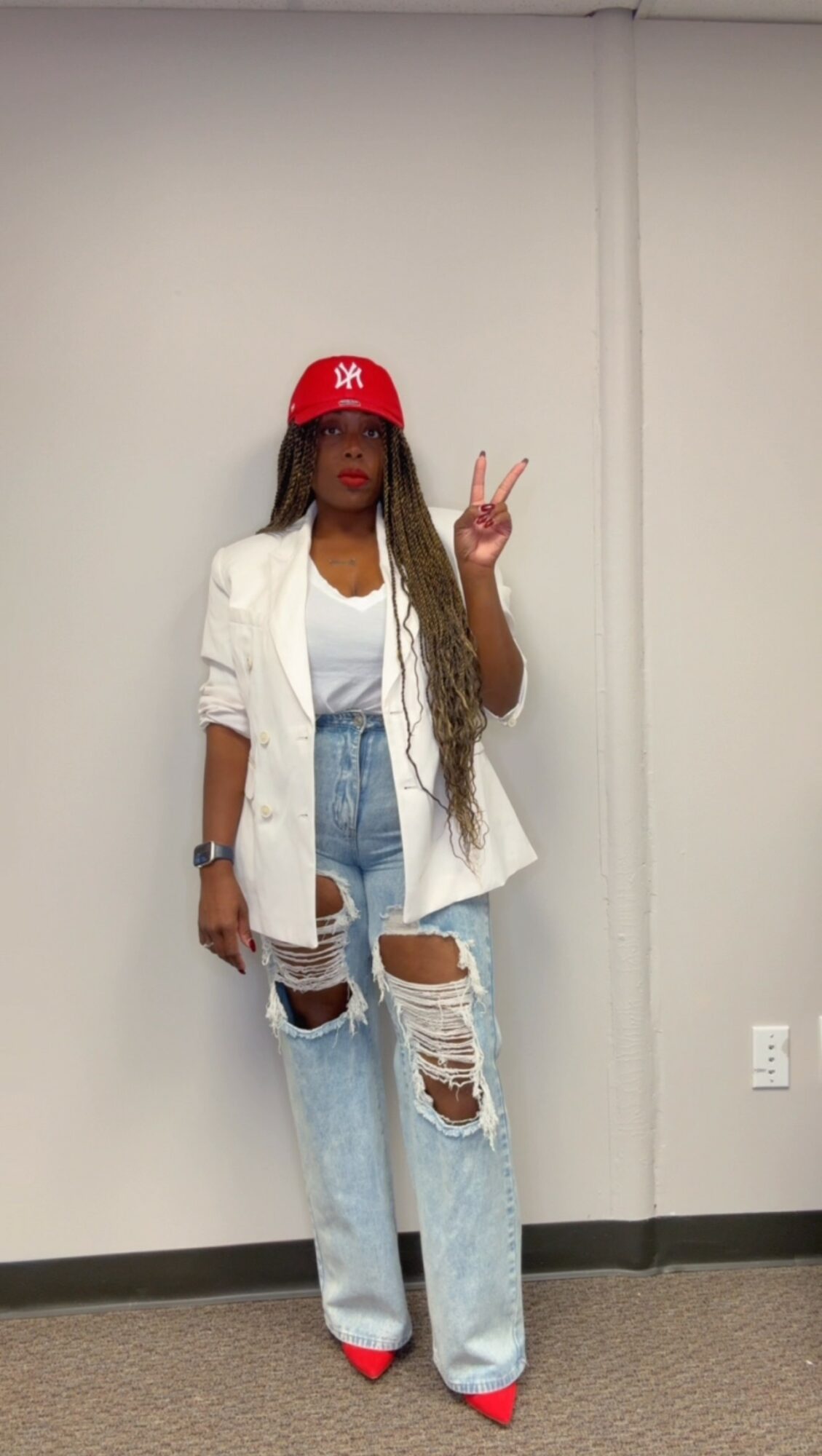Person with long hair wearing a red cap, white blazer, ripped jeans, and red shoes, making a peace sign.
