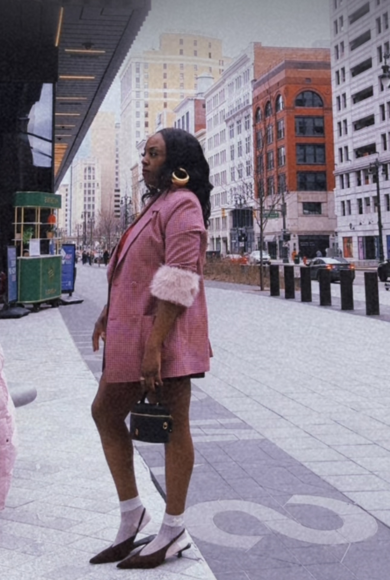Woman standing on city sidewalk with tall buildings in background, wearing pink coat, white shoes, and large earrings.