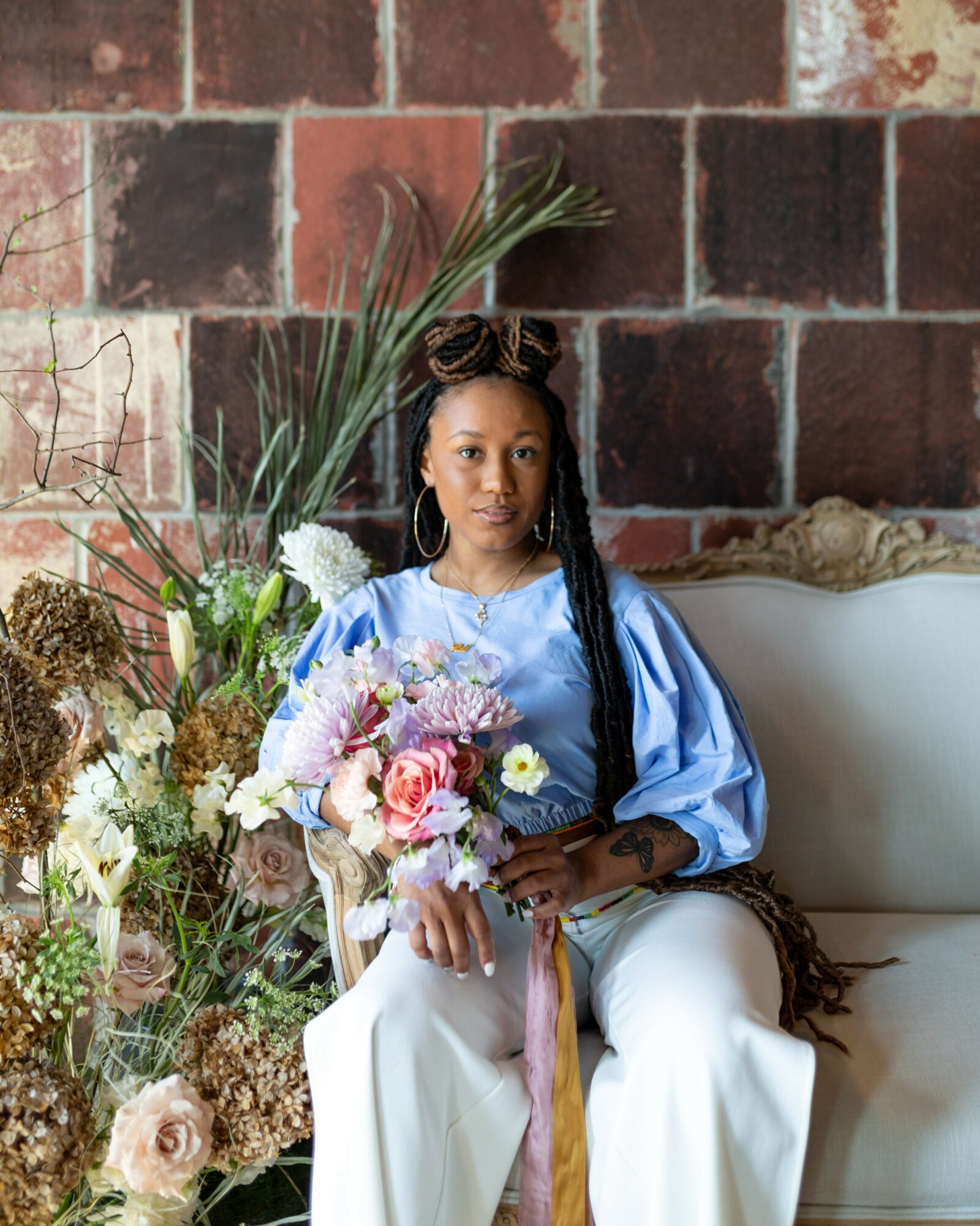 Young woman with long braided hair sitting on a white sofa, holding a bouquet of flowers, surrounded by floral arrangements, against a brick wall.