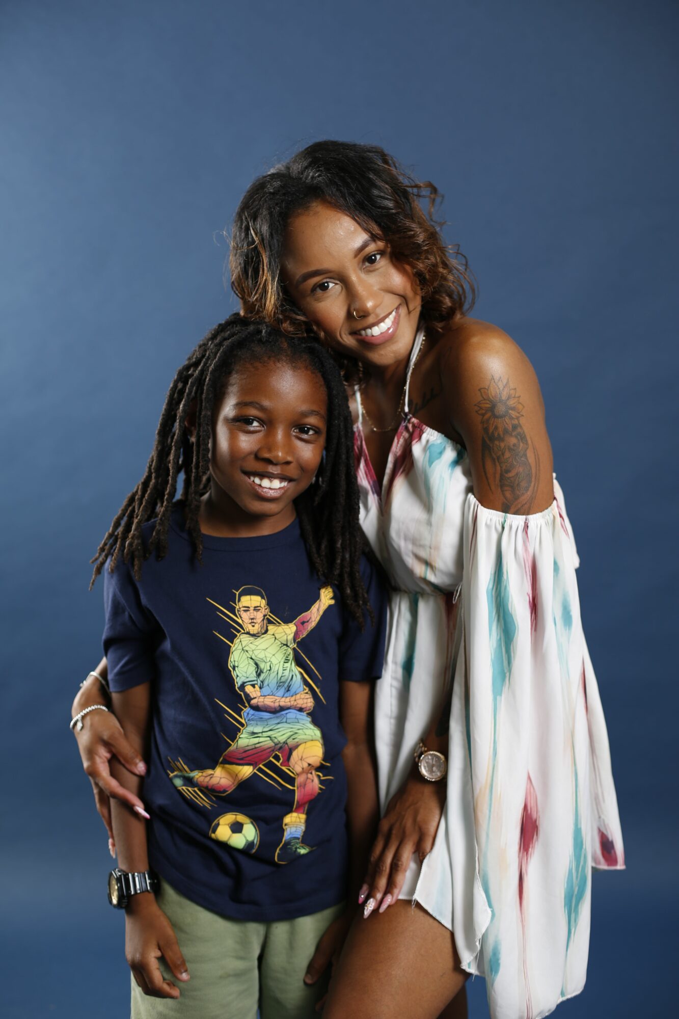 Two women smiling, one with dreadlocks and the other with curly hair, standing close together against a blue background.