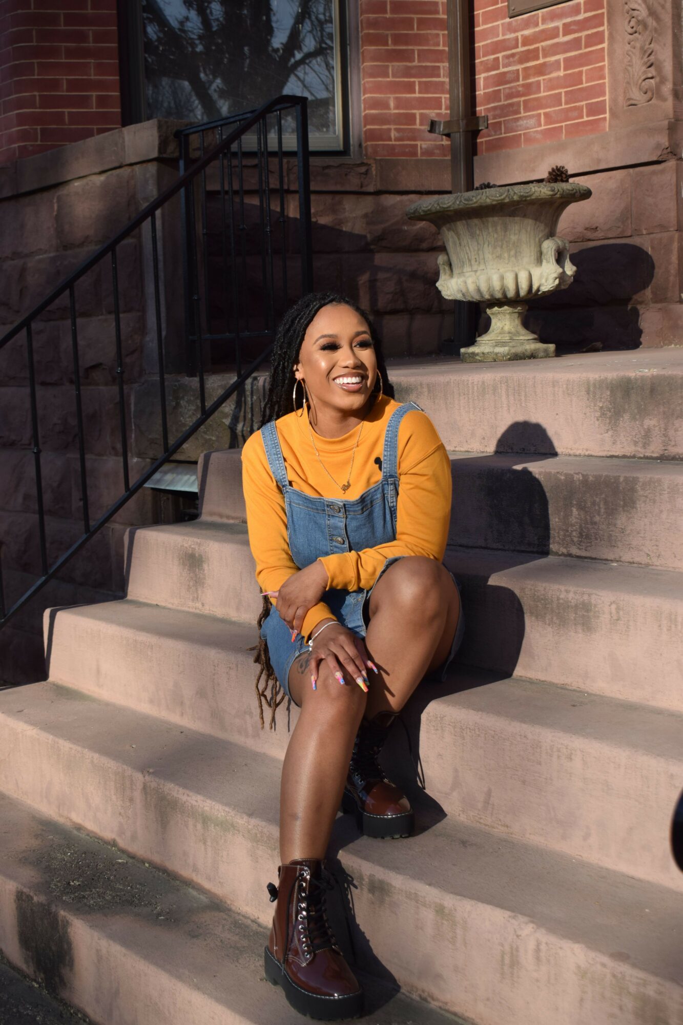 Woman sitting on stairs outside, wearing yellow jacket, black shorts, and boots, smiling at camera.
