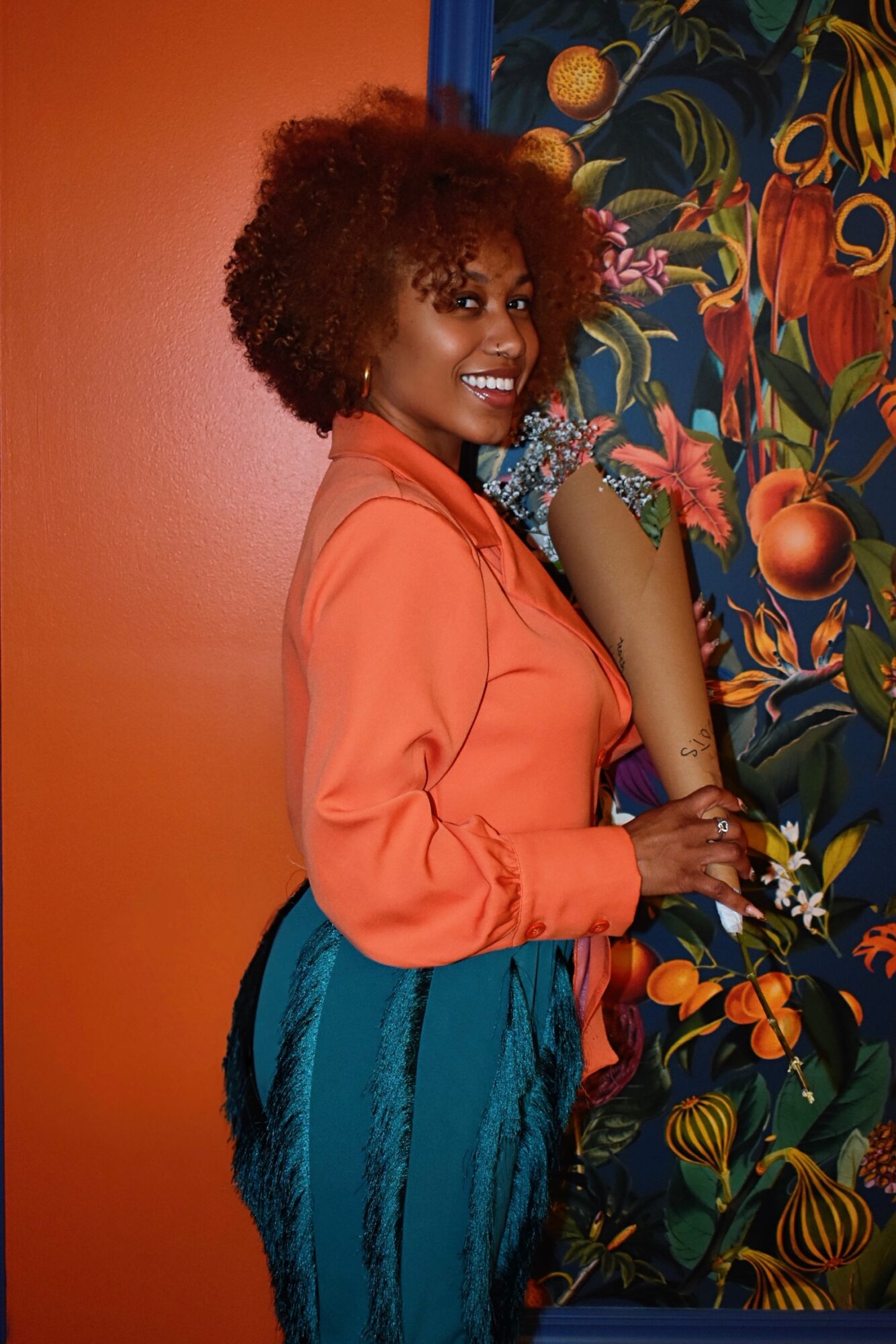Smiling woman with curly hair in an orange blouse and teal pants standing next to a colorful floral background.