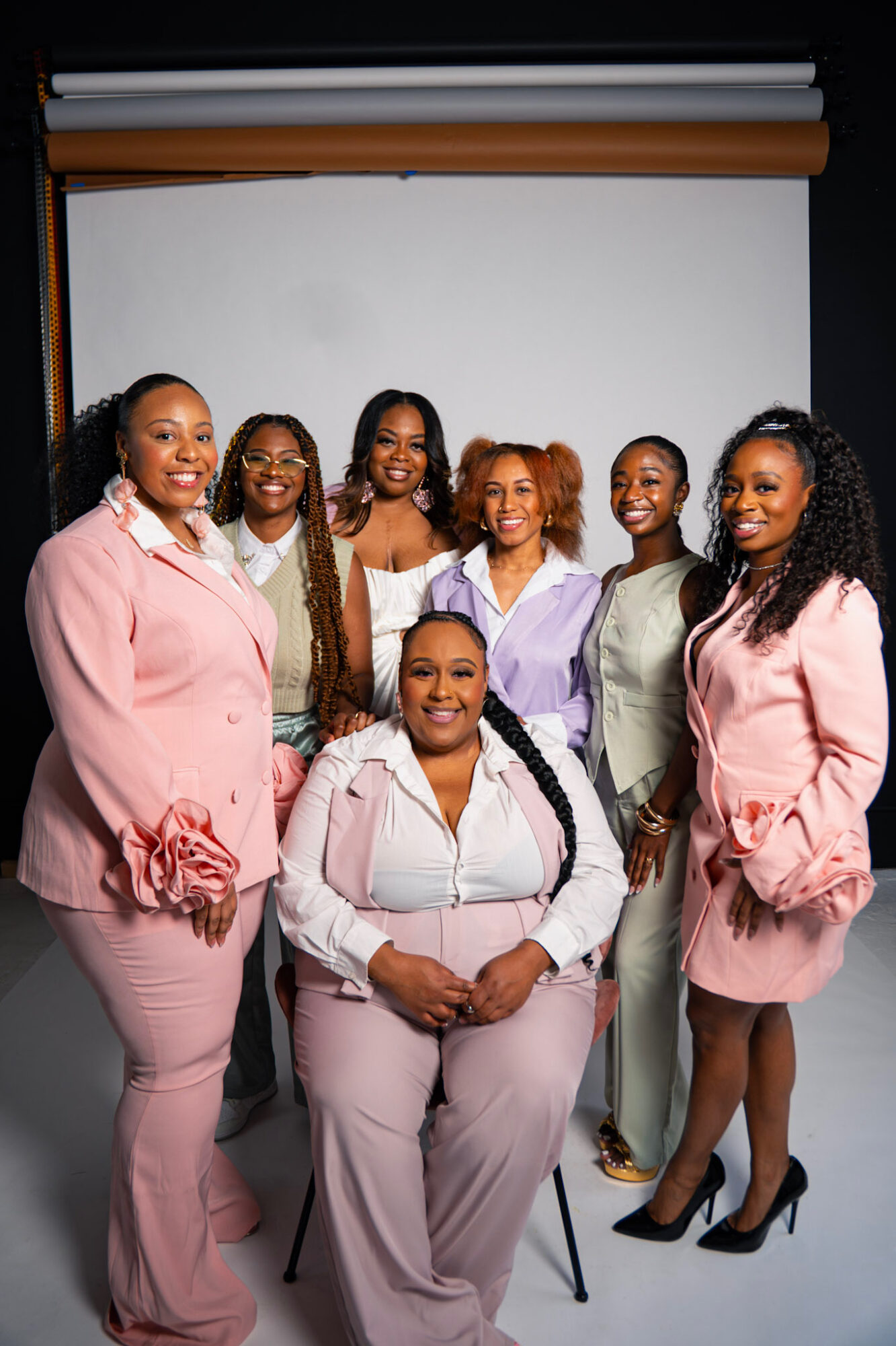 Group of seven women in pastel-colored outfits smiling in front of a white backdrop.