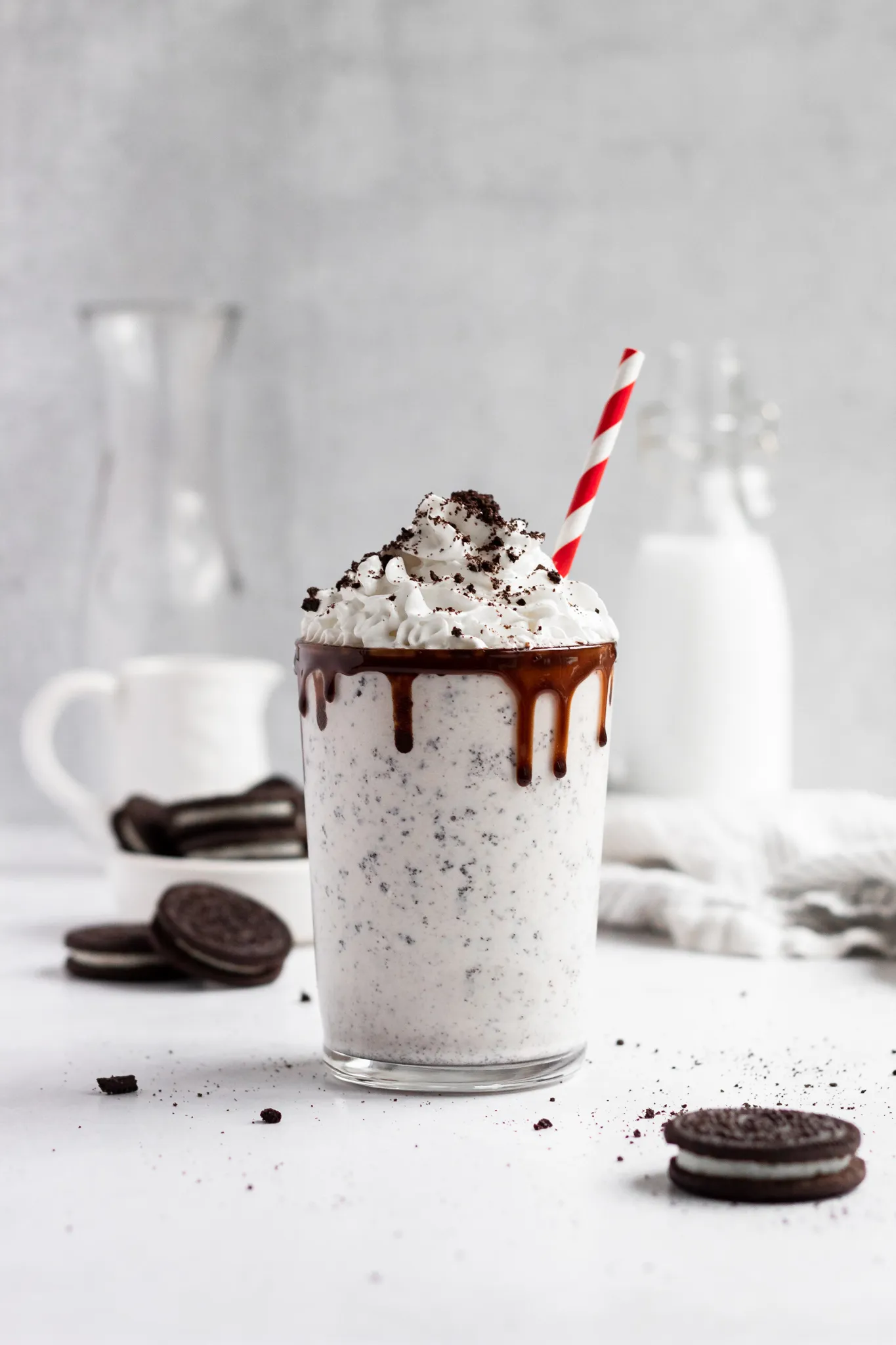 Milkshake topped with whipped cream and chocolate, with a striped straw, surrounded by cookies on a white surface.