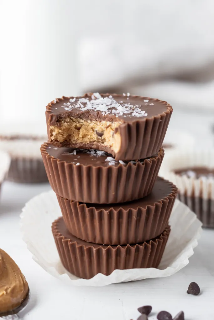 Stack of four chocolate cups with one bitten revealing a nut filling, on white paper, with chocolate chips nearby.