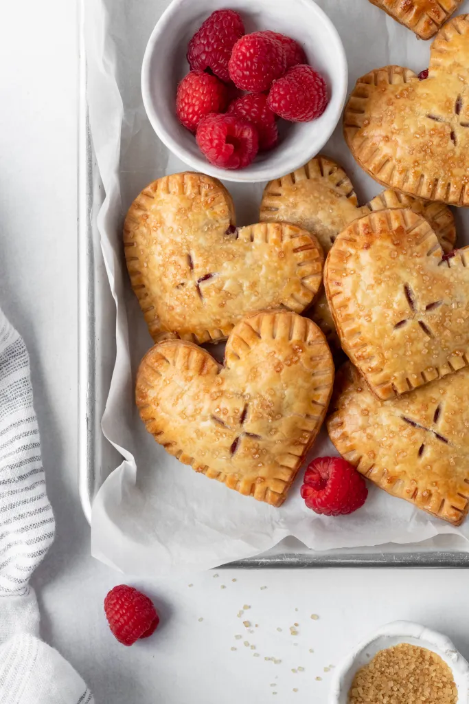 Heart-shaped pastries on white paper with a bowl of raspberries and a single raspberry nearby.