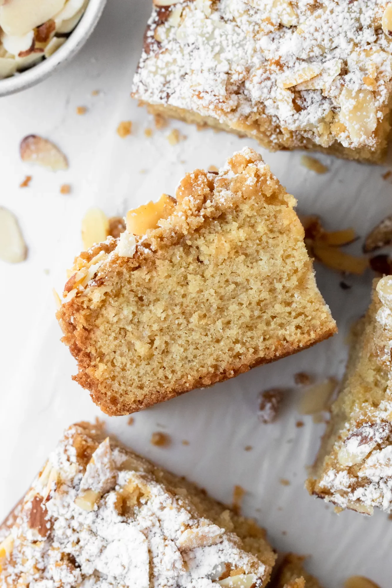 Slice of cake with powdered sugar and nuts on top, on a white surface with crumbs around.