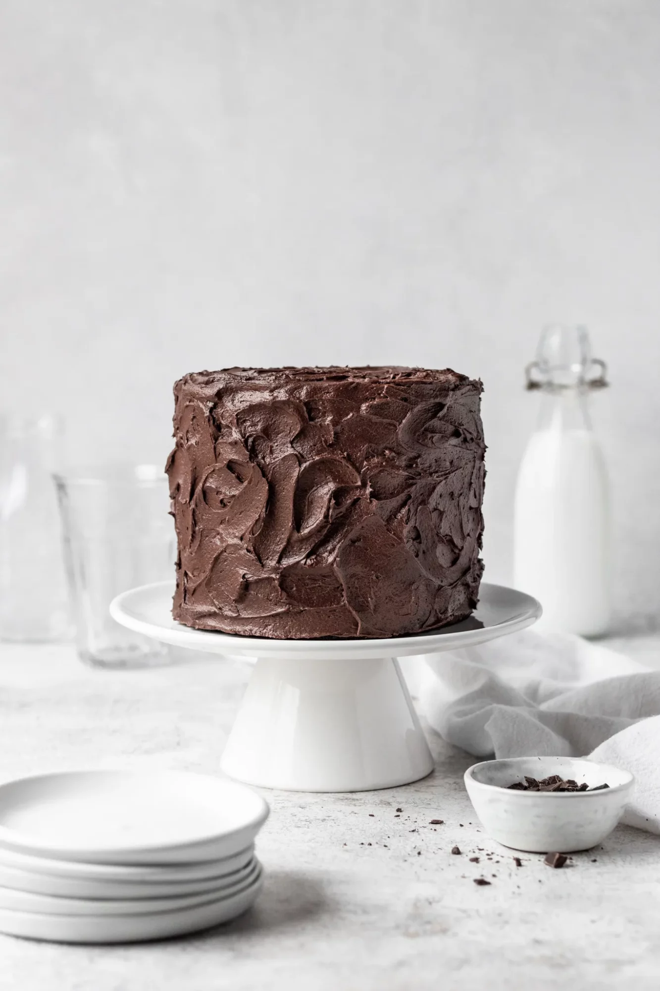 Chocolate cake on a white cake stand with plates and ingredients around, against a light background.