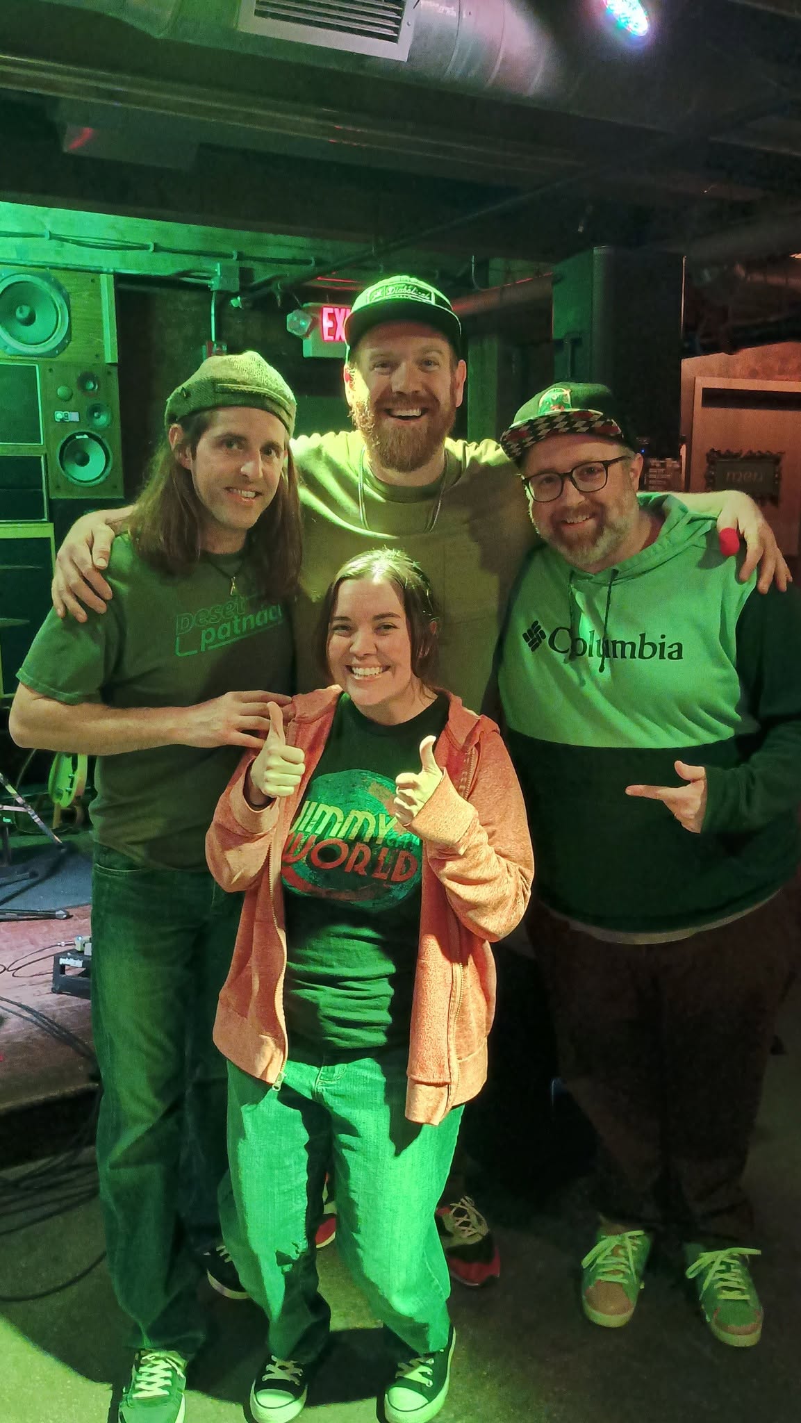 Four people smiling and posing together in a music venue, with speakers and green lighting in the background.