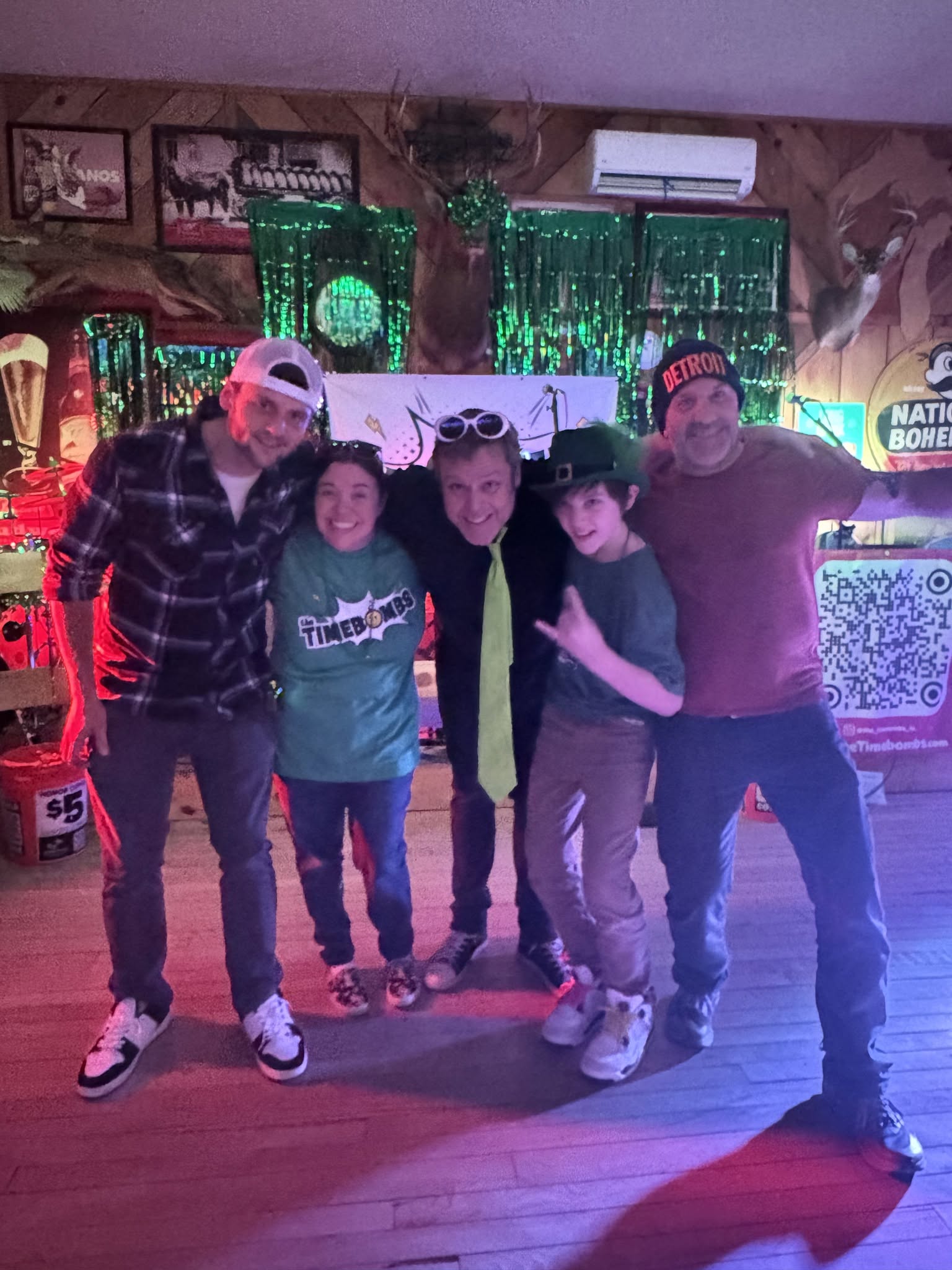 Four people standing together indoors, smiling, with neon green decorations and a QR code in the background.
