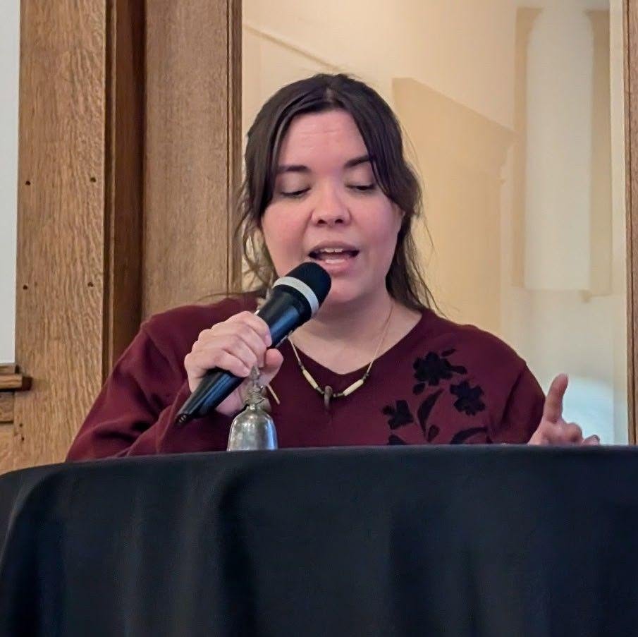 Woman speaking into a microphone, eyes closed, wearing a maroon top with black floral design, indoors.