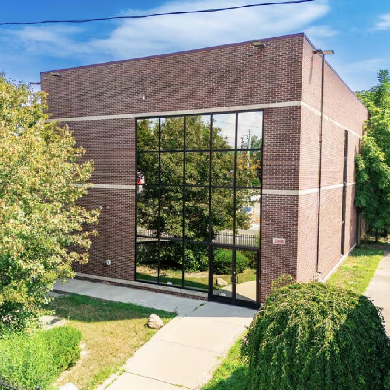 Brick building with large glass windows reflecting trees, surrounded by greenery and a sidewalk.
