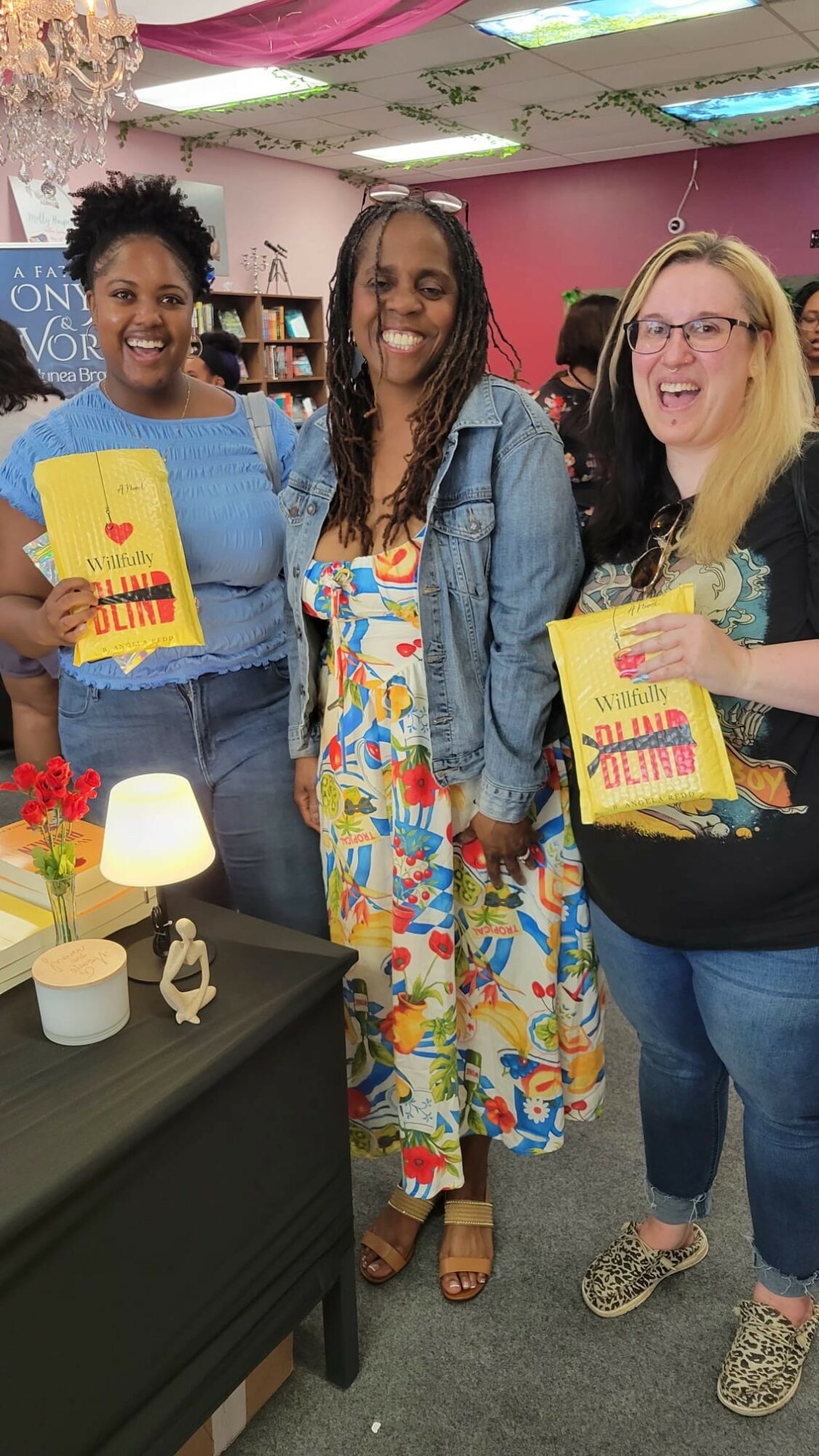 Three women standing together indoors, smiling, holding yellow booklets, with a table and decorations in the background.