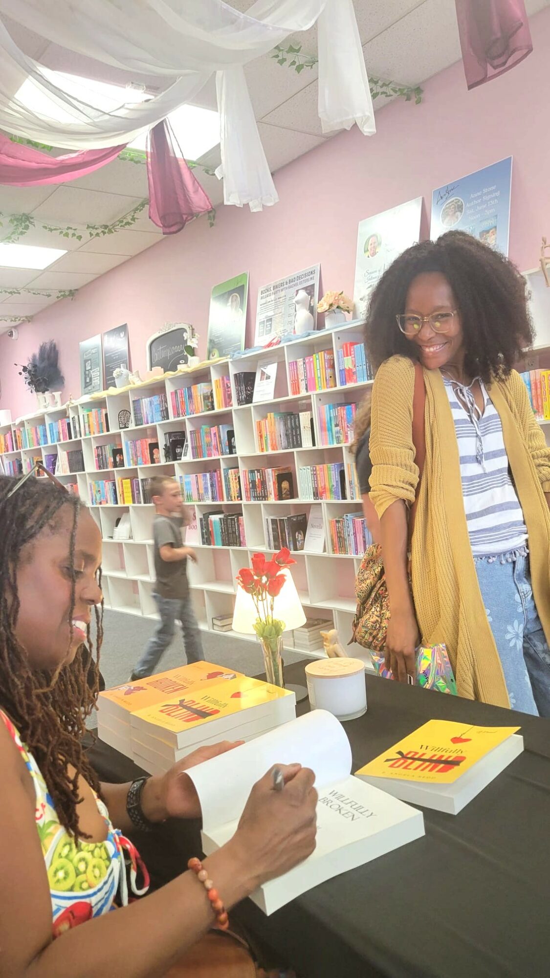 Two women at a book signing event in a bookstore, one signing a book, shelves with books in background.