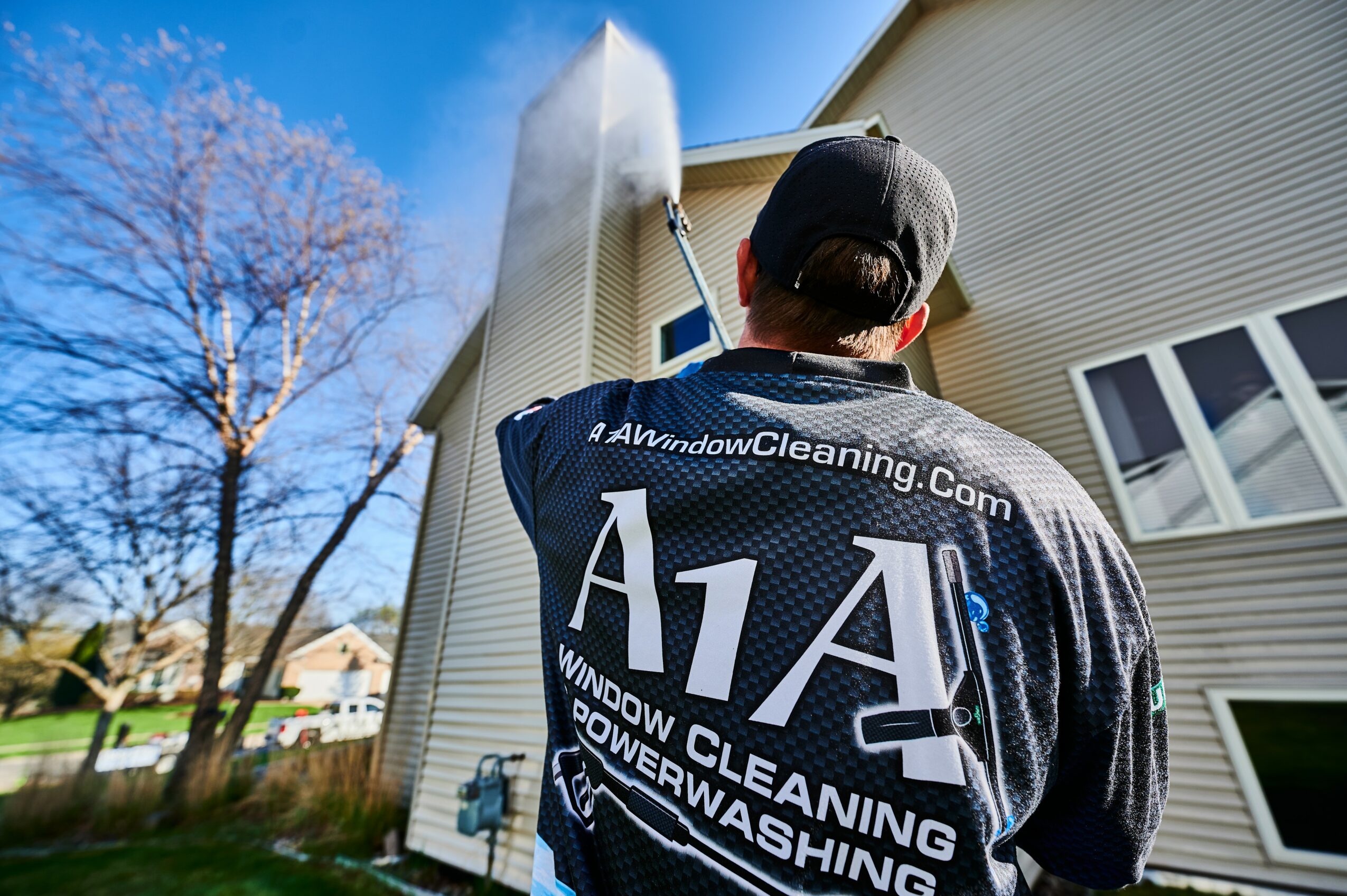 Person cleaning the exterior window of a house with a long-handled tool, wearing a dark cap and jacket, on a sunny day.